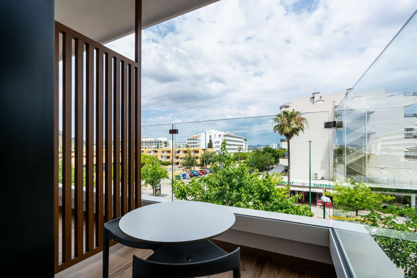 Blick vom Hotelbalkon mit Tisch und Glasgeländer auf grüne Bäume und umliegende Gebäude.