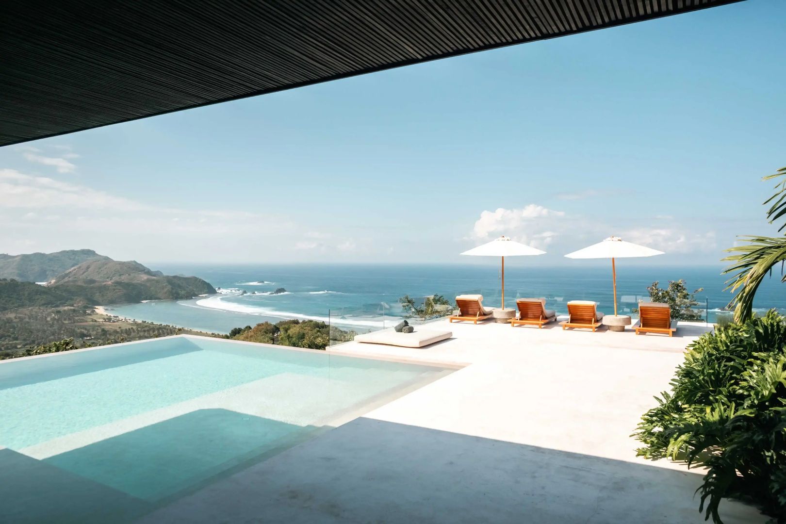 Infinity-Pool auf einer sonnigen Terrasse mit Liegestühlen und Blick auf die Meeresbucht.