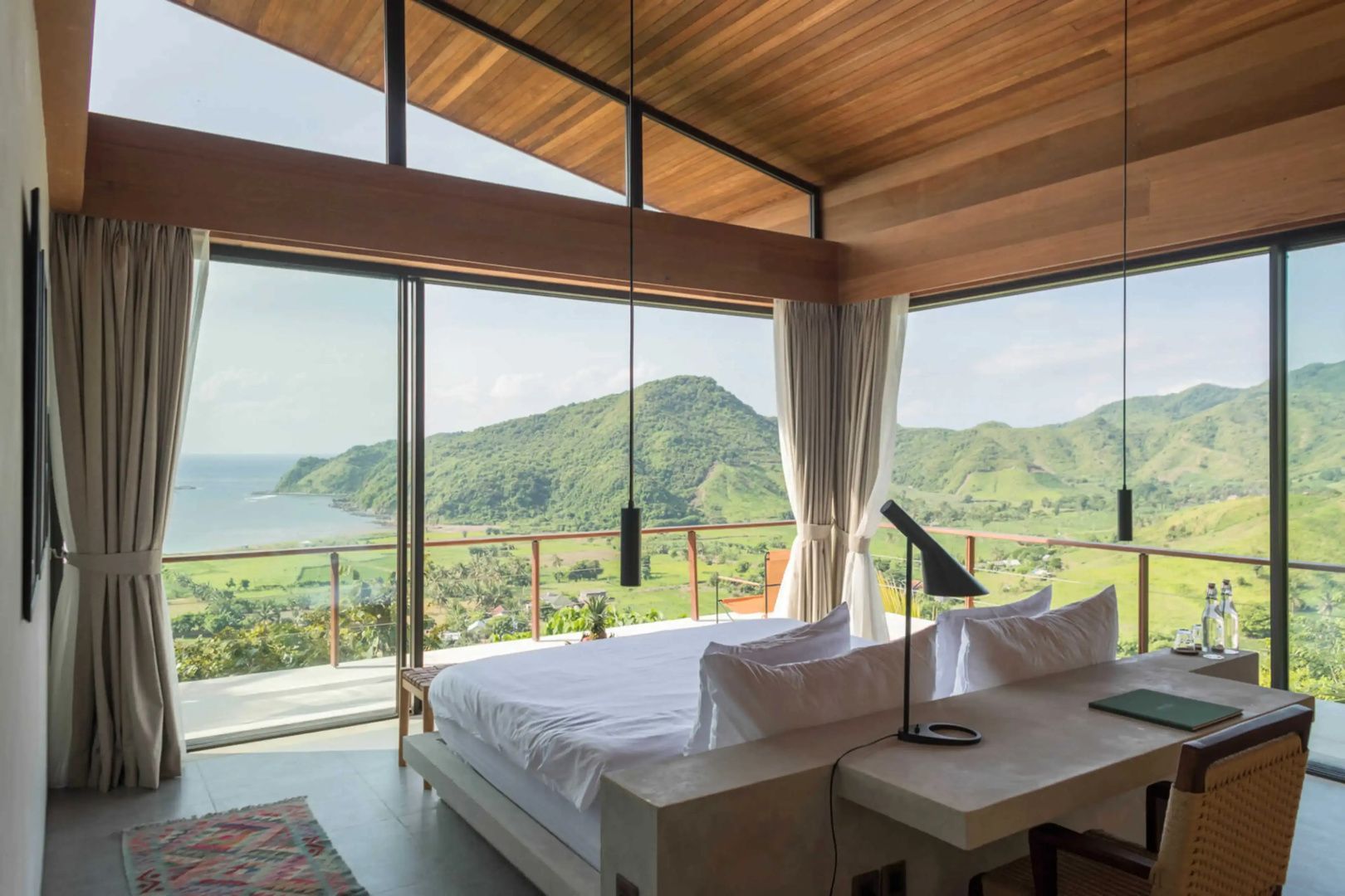 Schlafzimmer mit großen Panoramafenstern und Blick auf die grünen Hügel und das Meer.