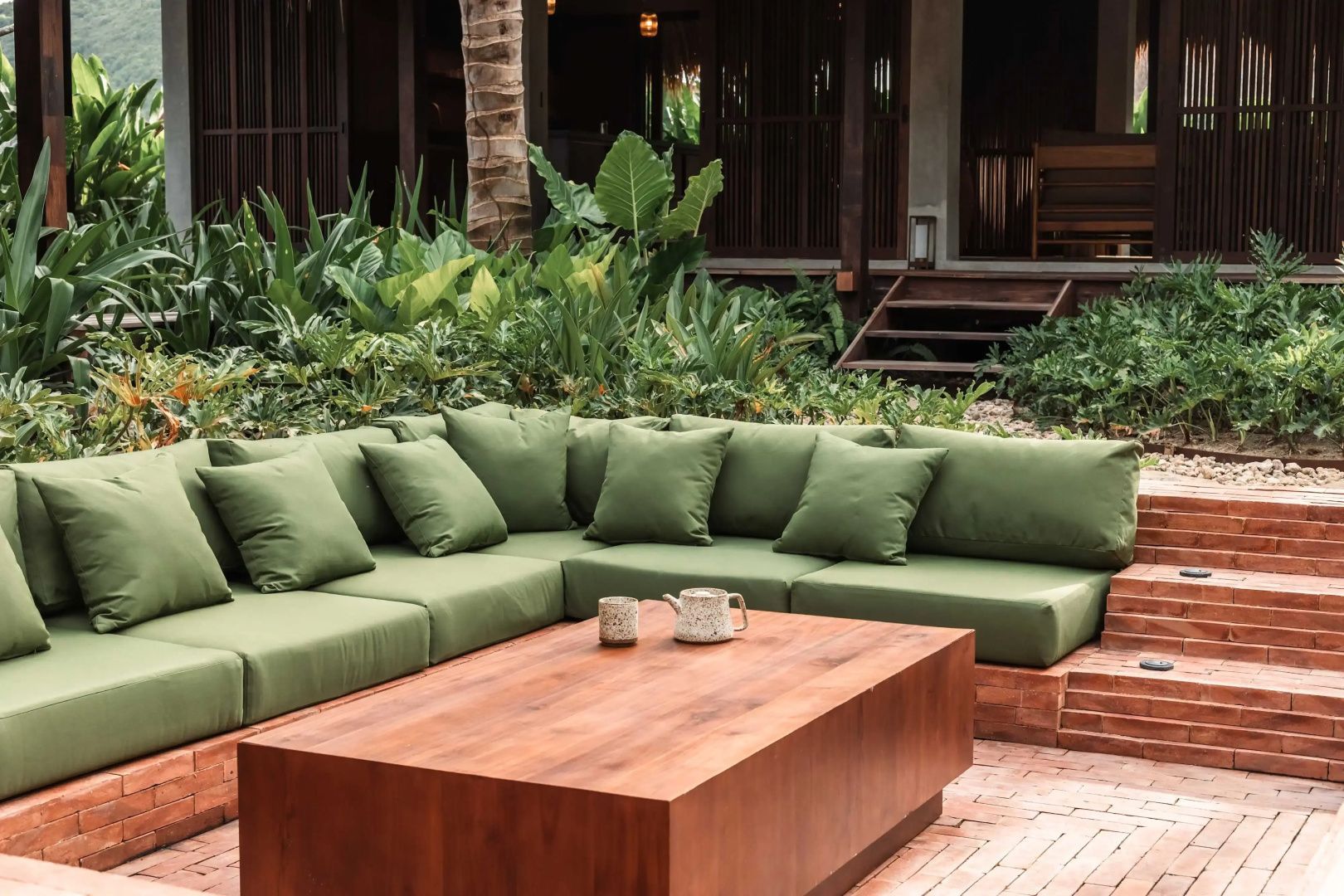 Grüne Couchgarnitur auf einer Terrasse im Garten neben tropischen Pflanzen.