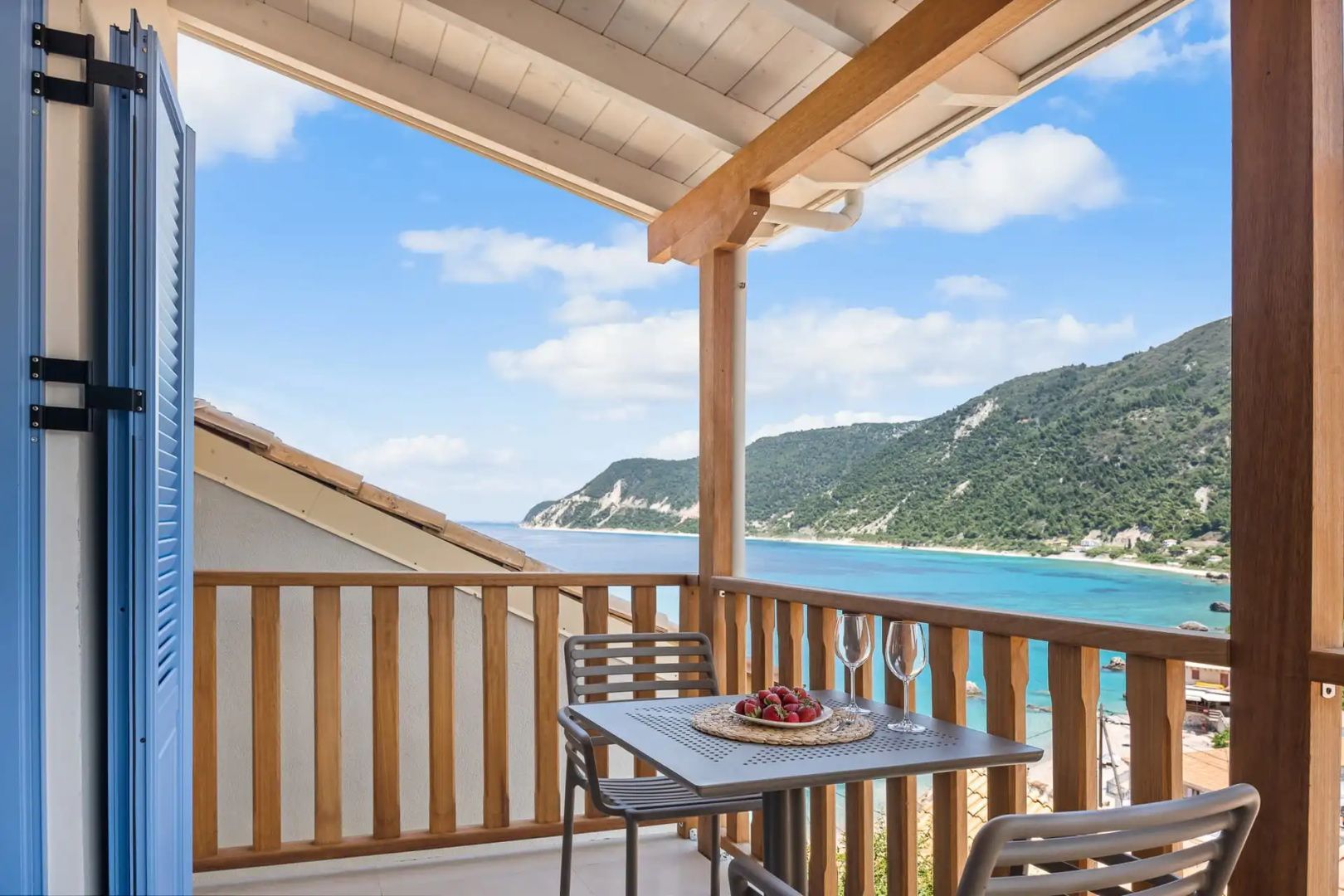 Balkon mit Holzgeländer, Tisch und Stühlen sowie Ausblick auf das Meer und grüne Berge.