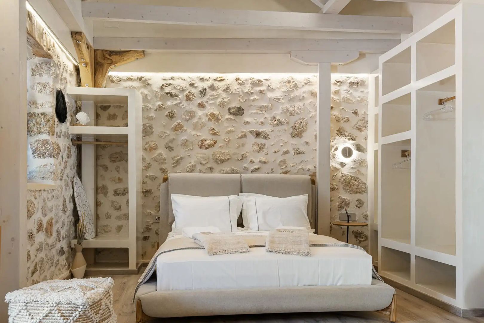 Schlafzimmer mit Doppelbett, beleuchteter Natursteinwand, Holzbalken und hellen Einbauregalen.