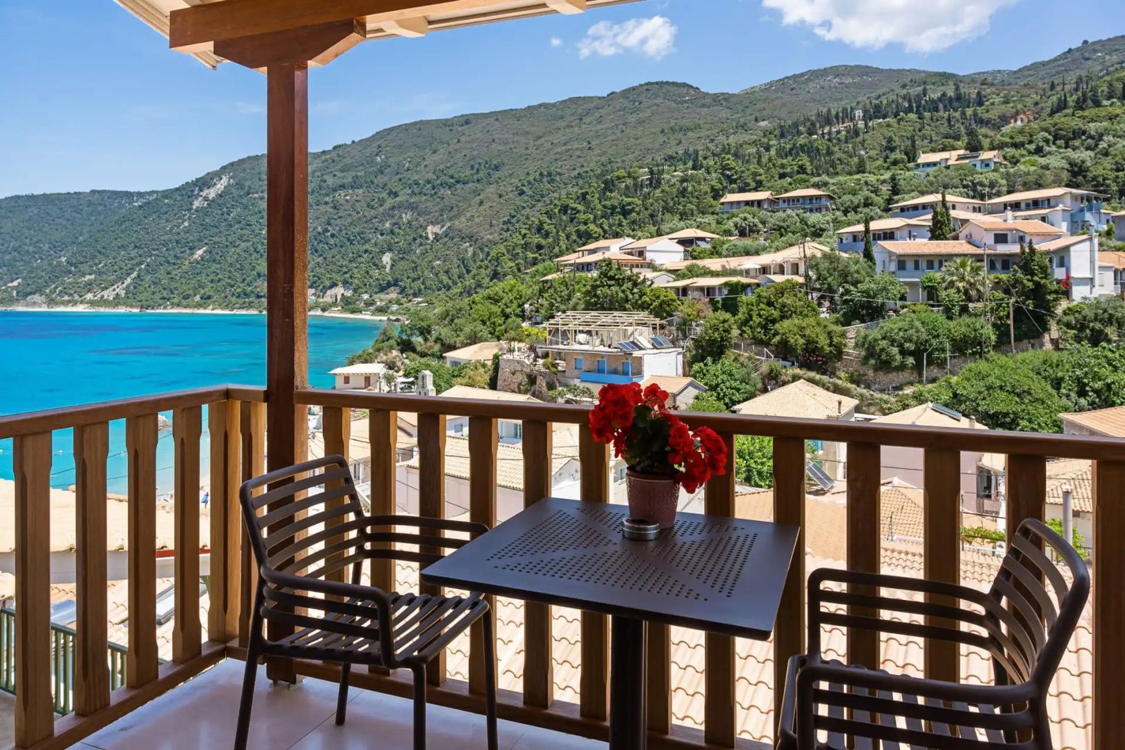 Balkon mit Tisch, Stühlen und Blumen, Blick auf ein Küstendorf sowie das blaue Meer.