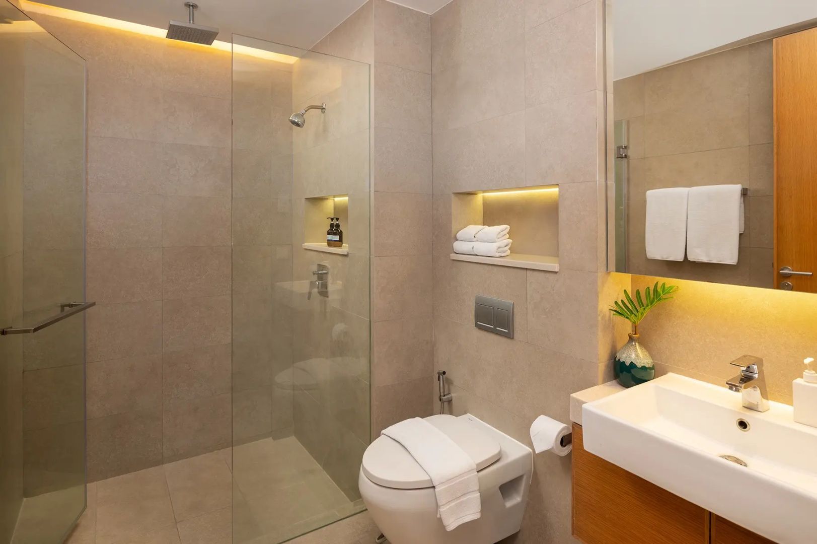 Badezimmer mit beigefarbenen Fliesen, begehbarer Glasdusche, Toilette und Waschtisch.