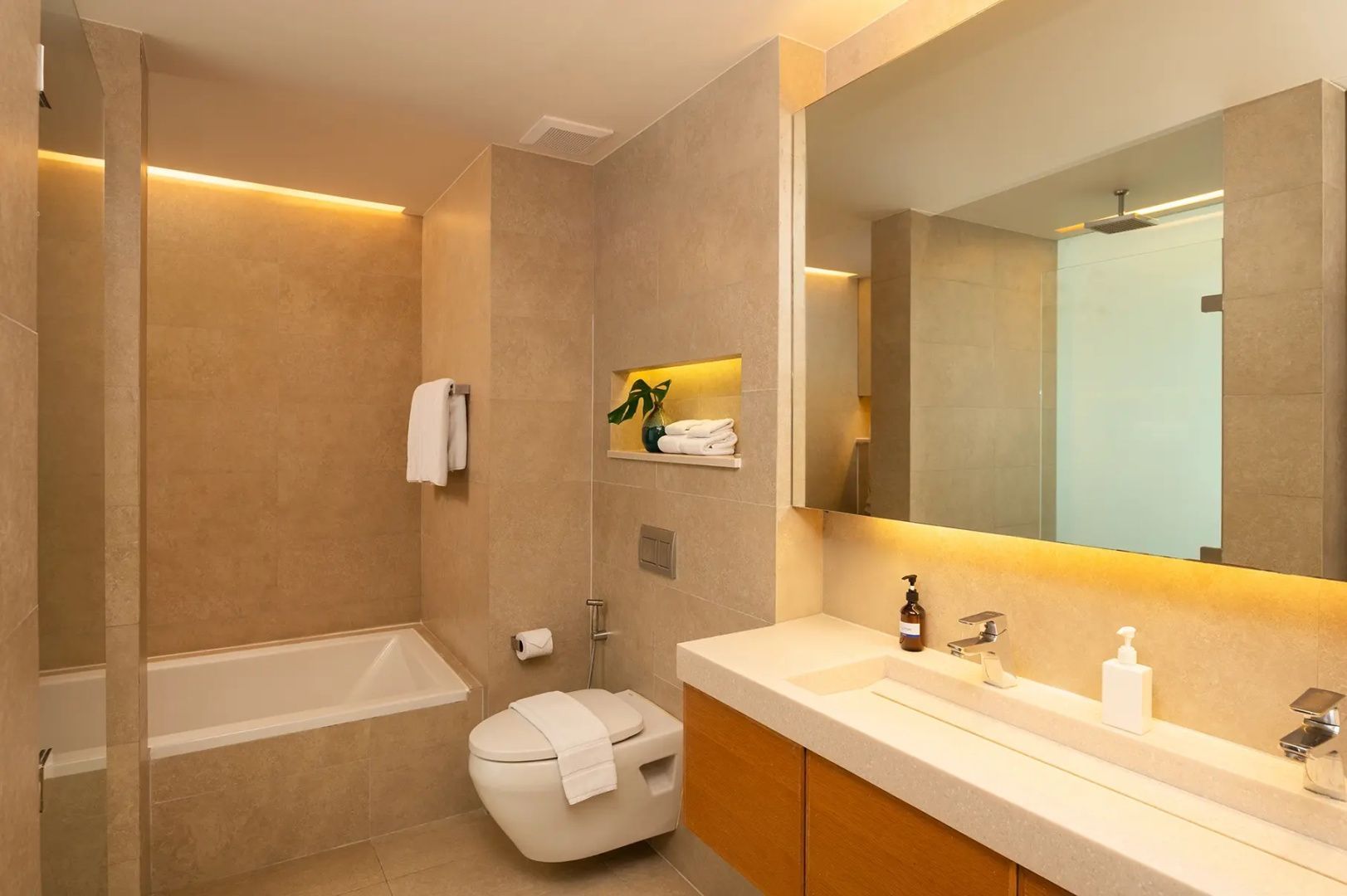 Beige gefliestes Badezimmer mit integrierter Badewanne, Wand WC und breitem Waschtisch mit Spiegel.