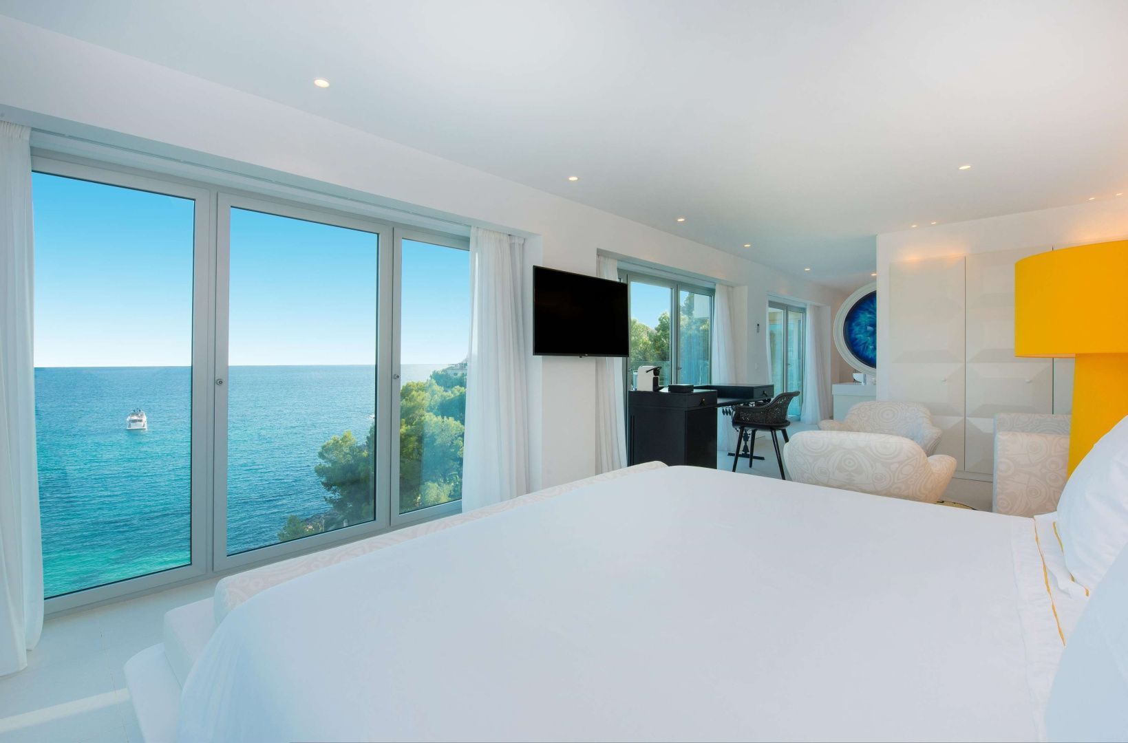 Blick aus dem Hotelzimmer durch große Fenster auf das blaue Meer und die grün bewachsene Küste.
