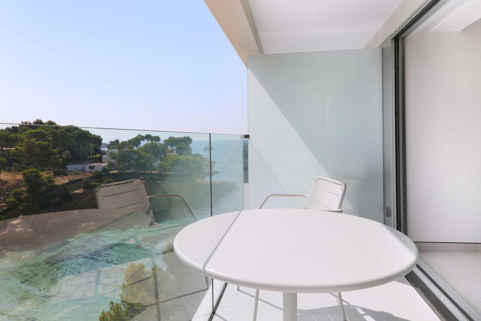 Balkon mit weißem Tisch, Stuhl, Glasgeländer und Blick auf Küste und Meer.