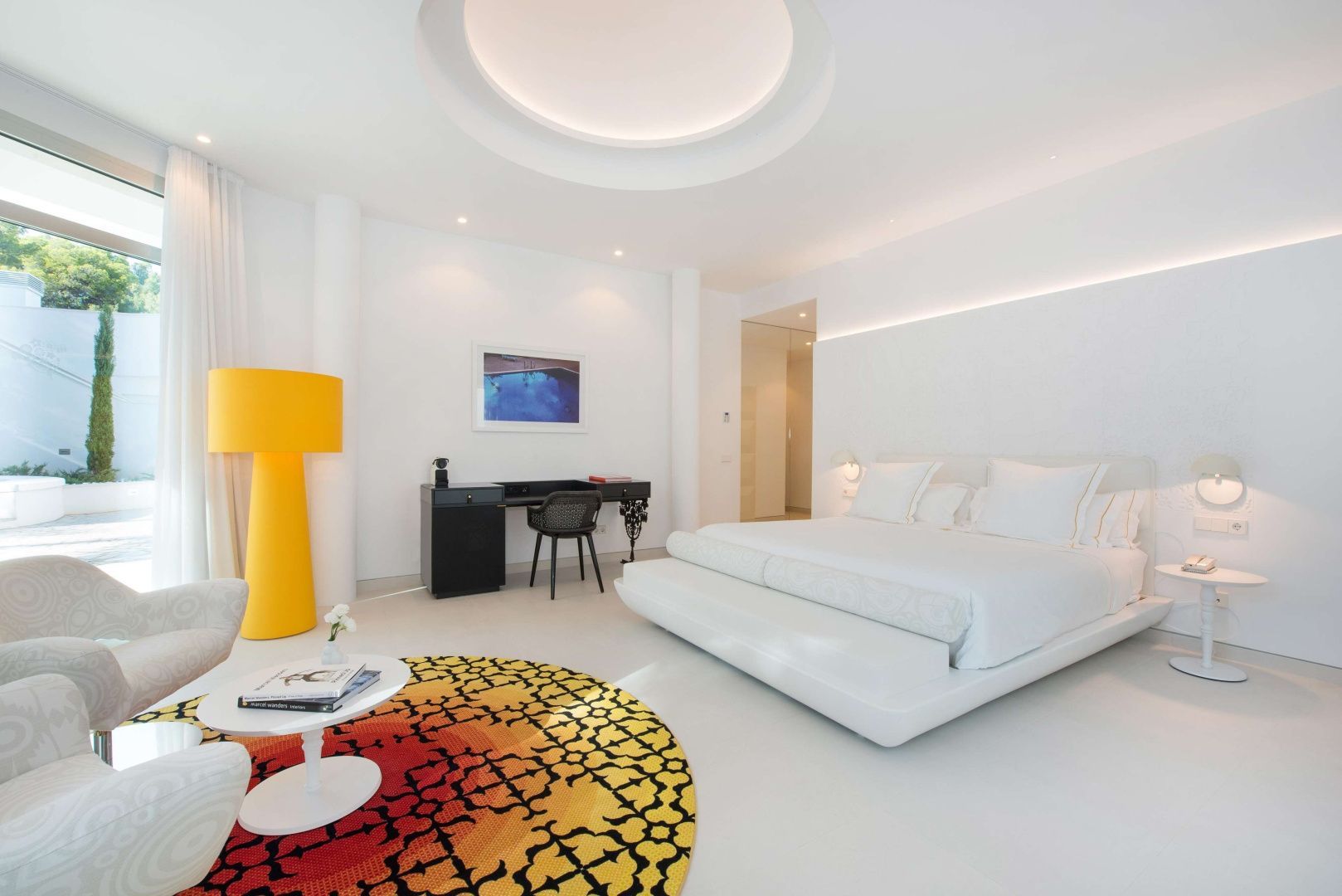 Modernes weißes Hotelzimmer mit großem Doppelbett, gelber Stehlampe und buntem rundem Teppich.