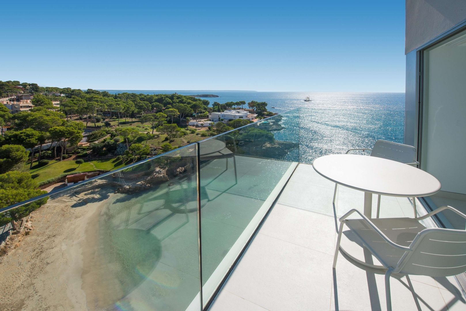 Blick von der Terrasse über eine Glasbrüstung auf den Strand, grüne Bäume und das offene Meer.