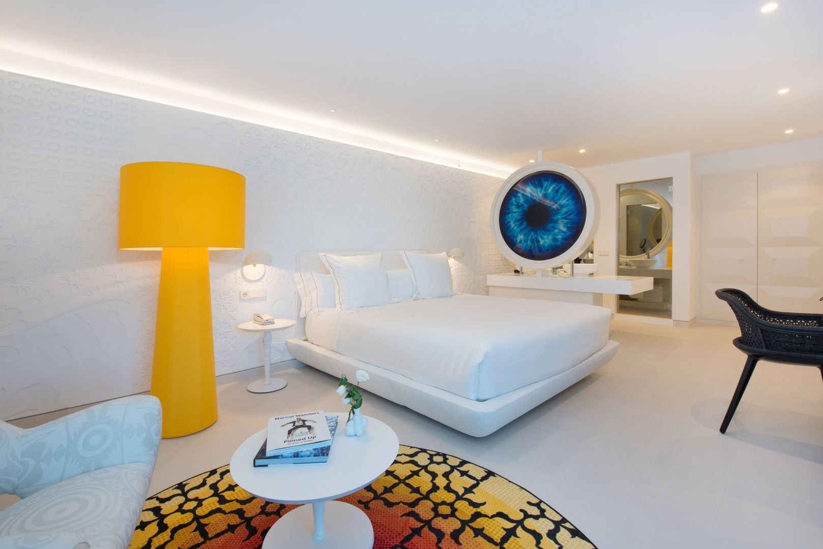 Helles Hotelzimmer mit weißem Doppelbett, gelber Stehlampe und Kunstwerk an der Wand.