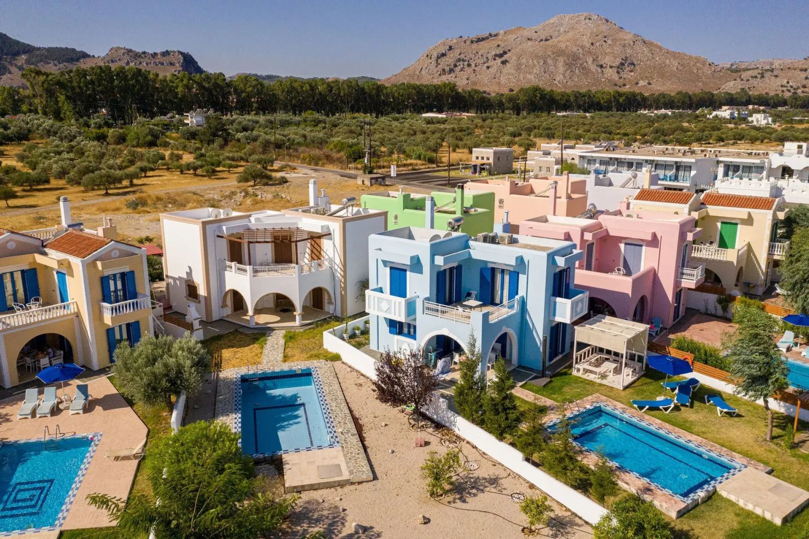 Farbenfrohe Ferienvillen mit privaten Pools vor einer Landschaft mit Bergen.