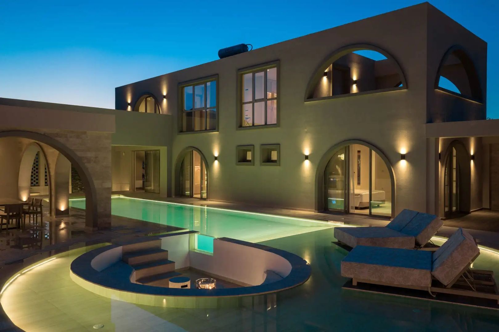 Beleuchtete Villa bei Dämmerung mit Swimmingpool, versenkter Sitzecke und Liegestühlen im Außenbereich.