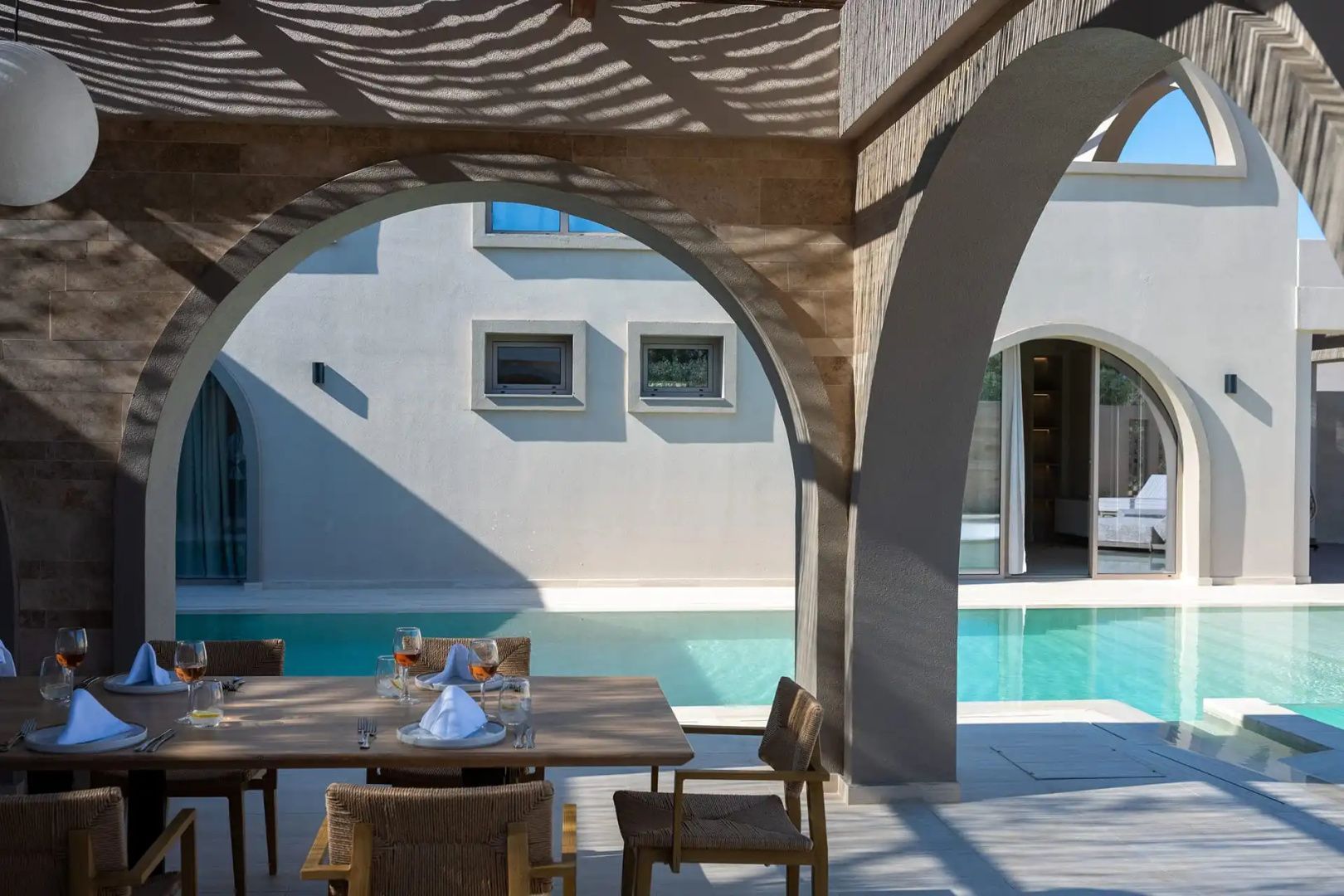 Gedeckter Tisch unter einer Pergola neben einem Pool und hellem Gebäudeteil.