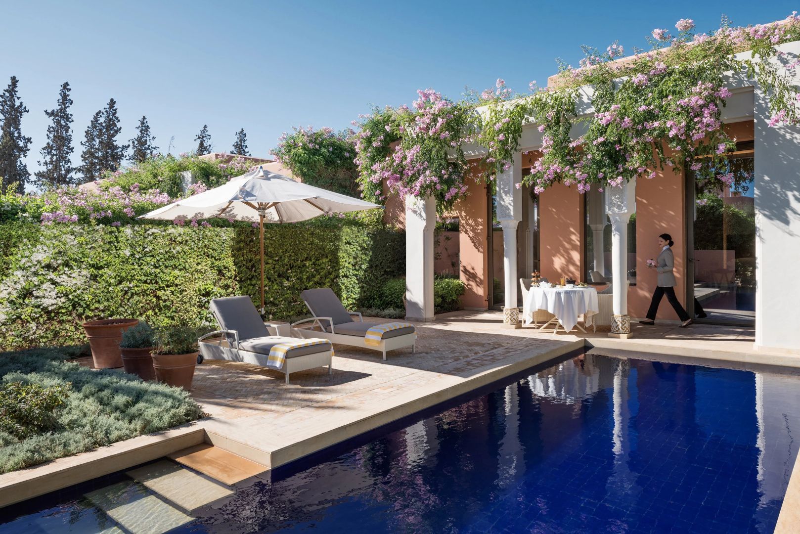 Sonnenterrasse am tiefblauen Pool mit zwei Liegestühlen, Sonnenschirm und blühenden Pflanzen.