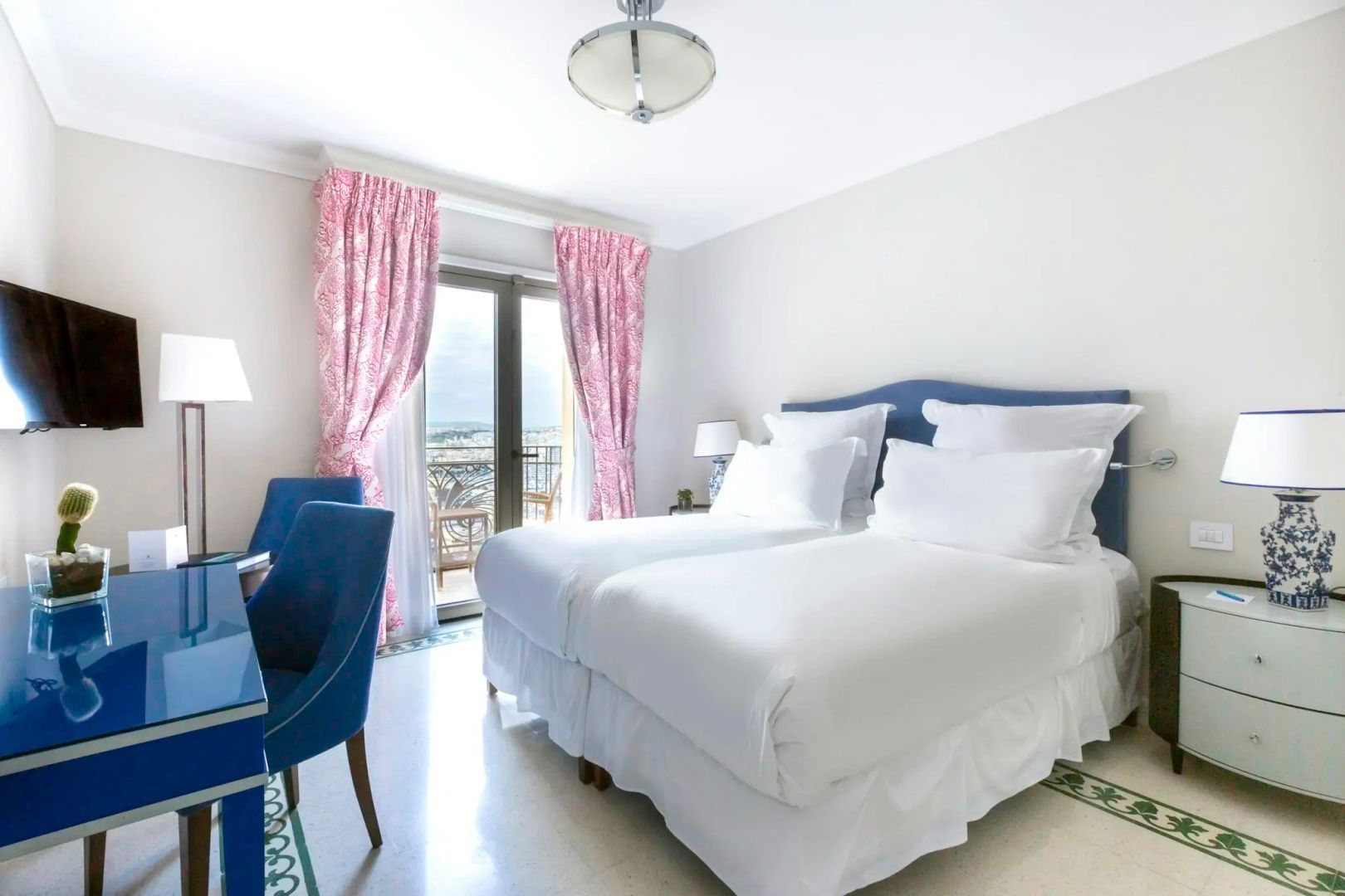 Helles Hotelzimmer mit zwei Betten, blauen Sesseln, rosa Vorhängen und Balkon.