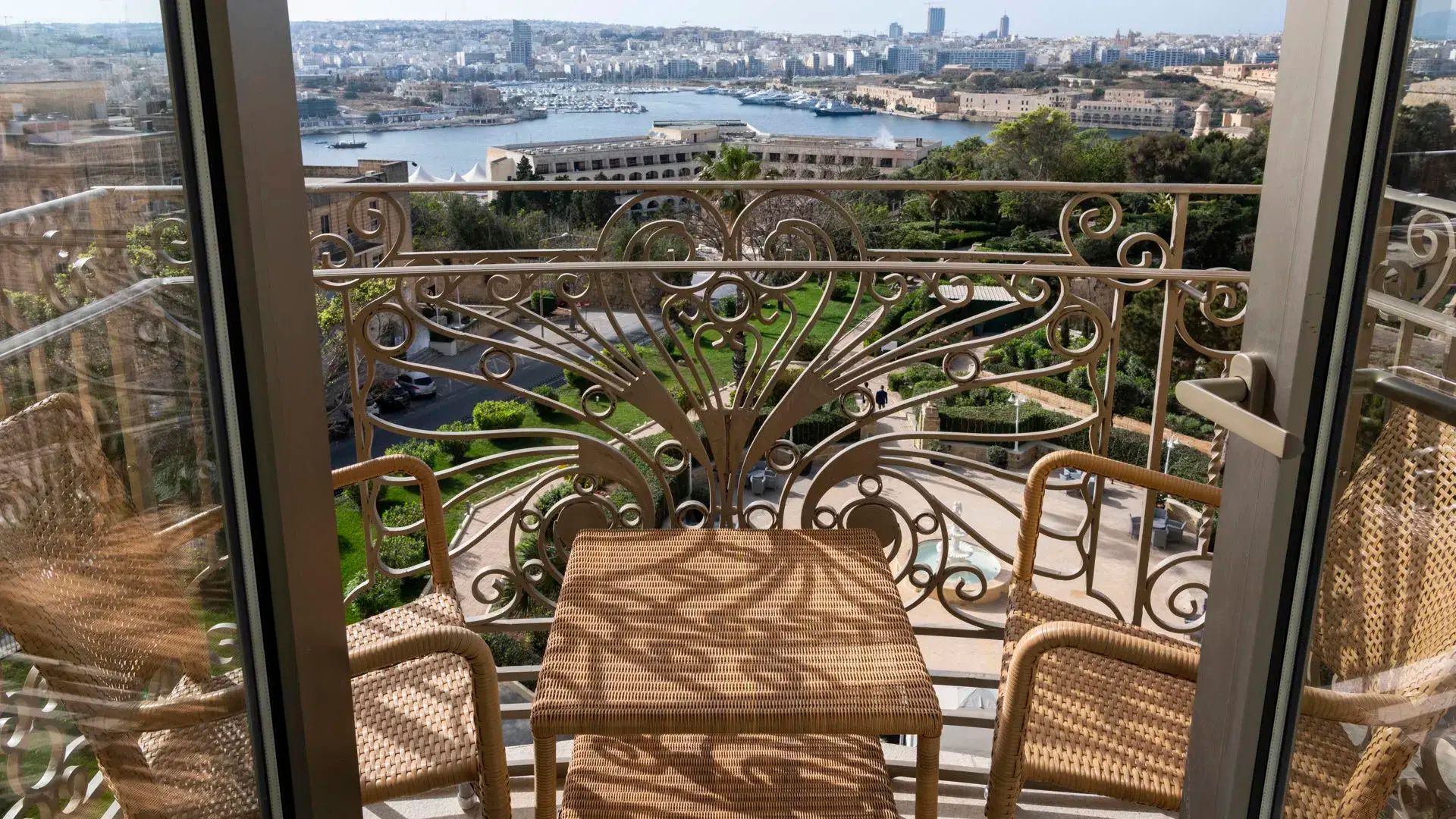Balkon mit Flechtstühlen und verschnörkeltem Geländer mit Blick auf den Garten und den Hafen.
