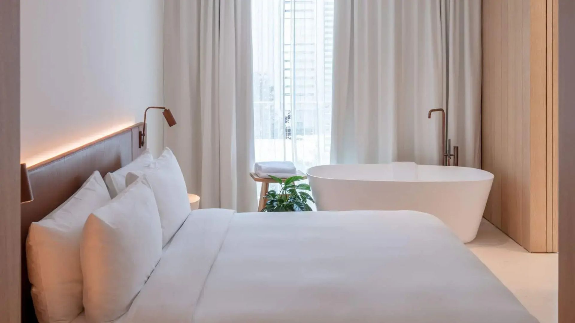 Helles Hotelzimmer mit weiß bezogenem Bett und freistehender Badewanne vor dem Fenster.