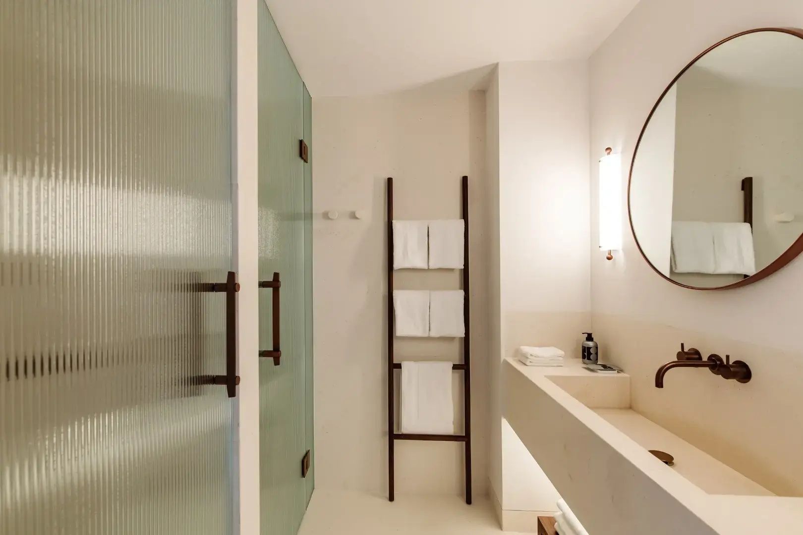 Badezimmer mit hellem Waschtisch, Handtuchleiter aus Holz und einer matten Glastür.