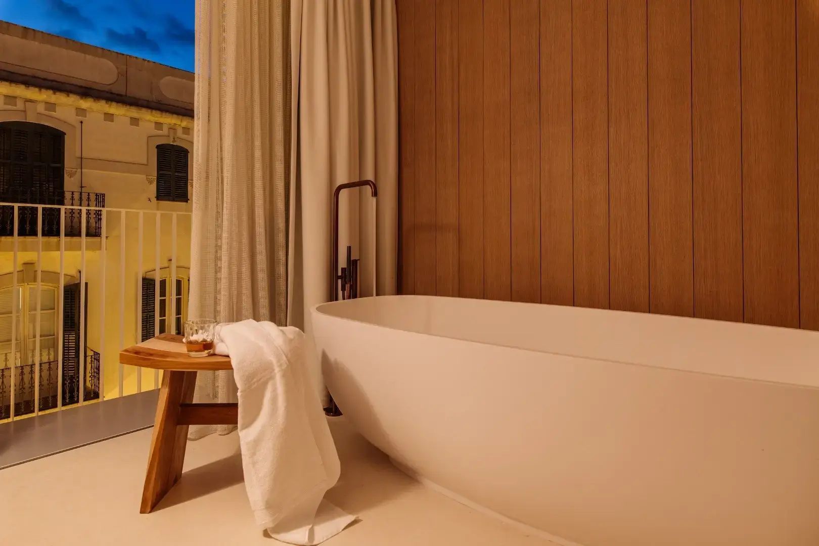 Freistehende weiße Badewanne vor einer Holzwand mit kleinem Hocker davor.