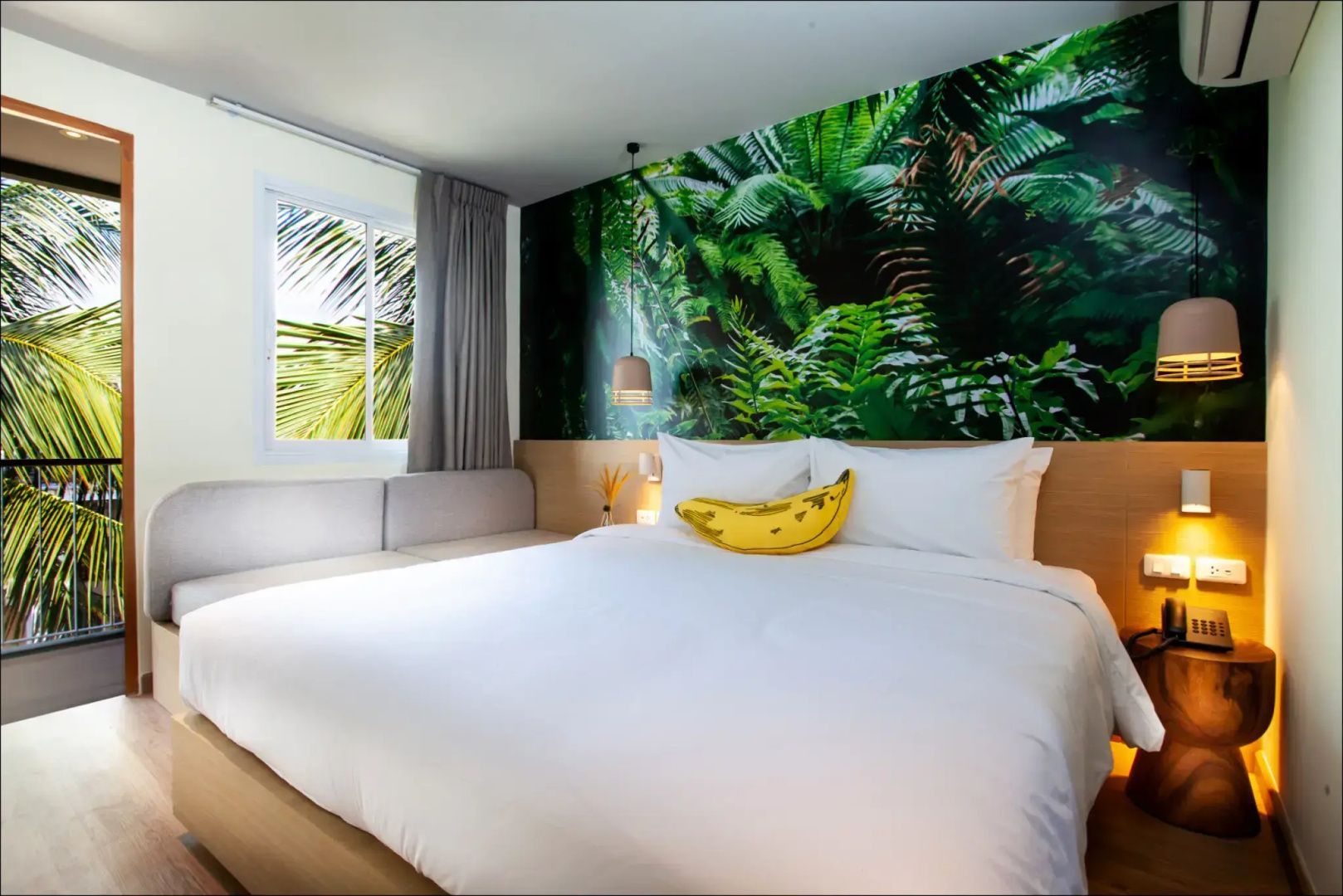 Doppelbett mit weißer Bettwäsche, gelbem Bananenkissen und tropischer Fototapete im Zimmer.