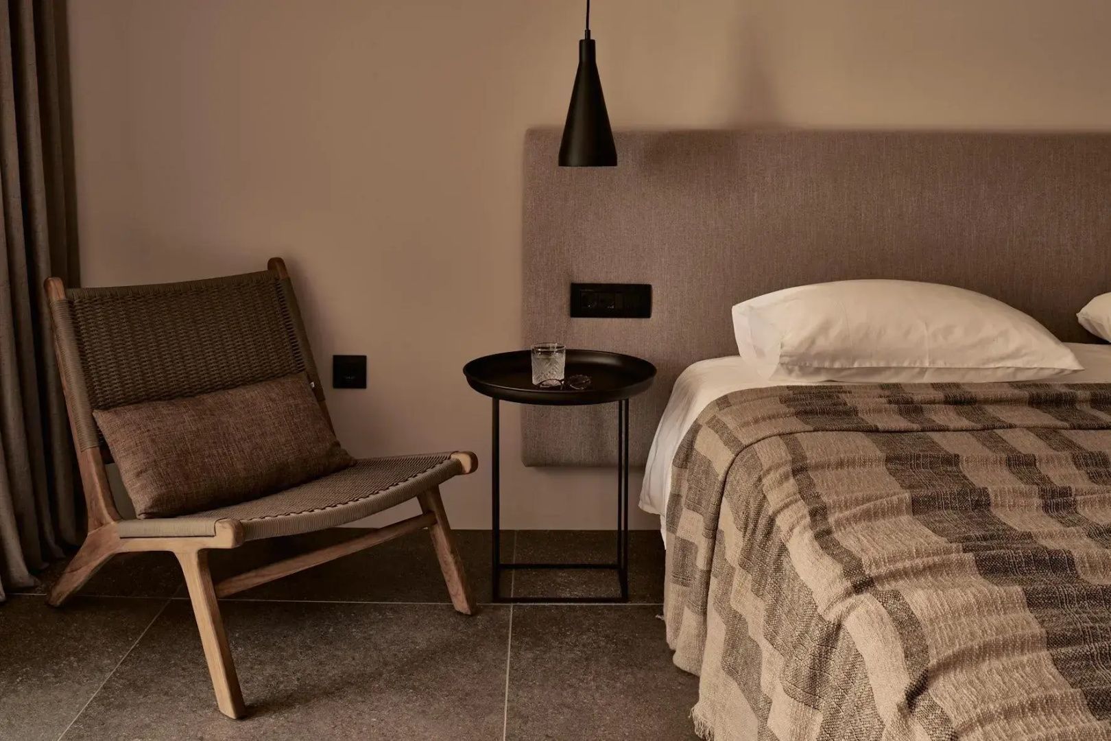 Sessel aus Geflecht und runder Nachttisch neben einem Bett mit karierter Decke.