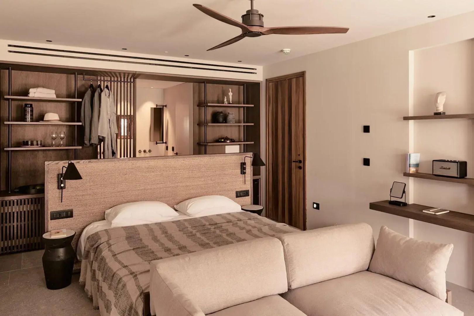 Hotelzimmer mit geordnetem Doppelbett, Sofa, Holzdeckenventilator und offenem Schrankbereich.