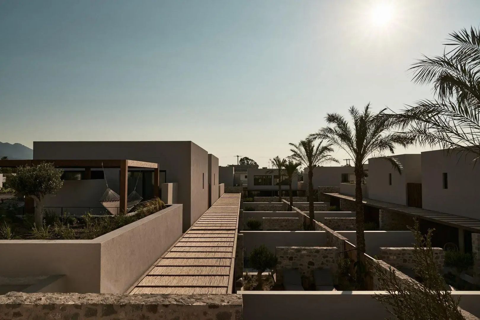 Modernes Hotelgebäude mit Pergola aus Holz, Natursteinmauern und hoch gewachsenen Palmen im Außenbereich.