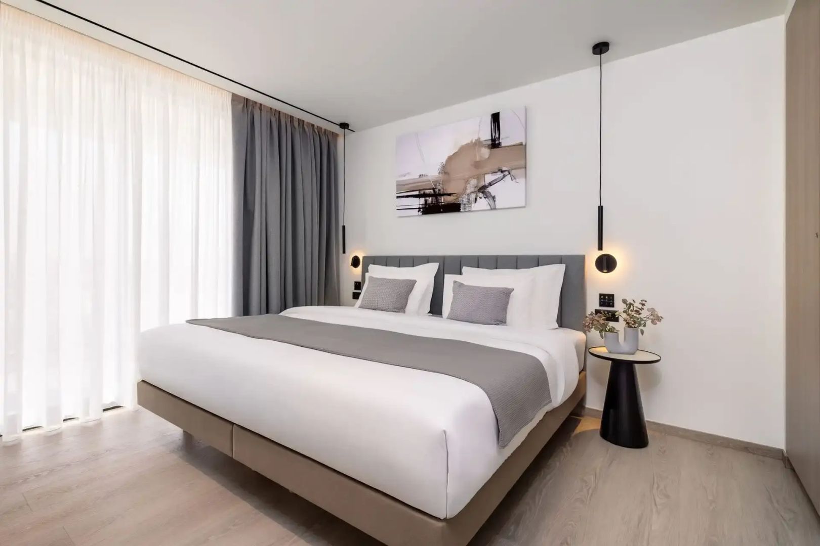 Doppelbett mit grauer Tagesdecke und gepolstertem Kopfteil in einem hellen Zimmer.