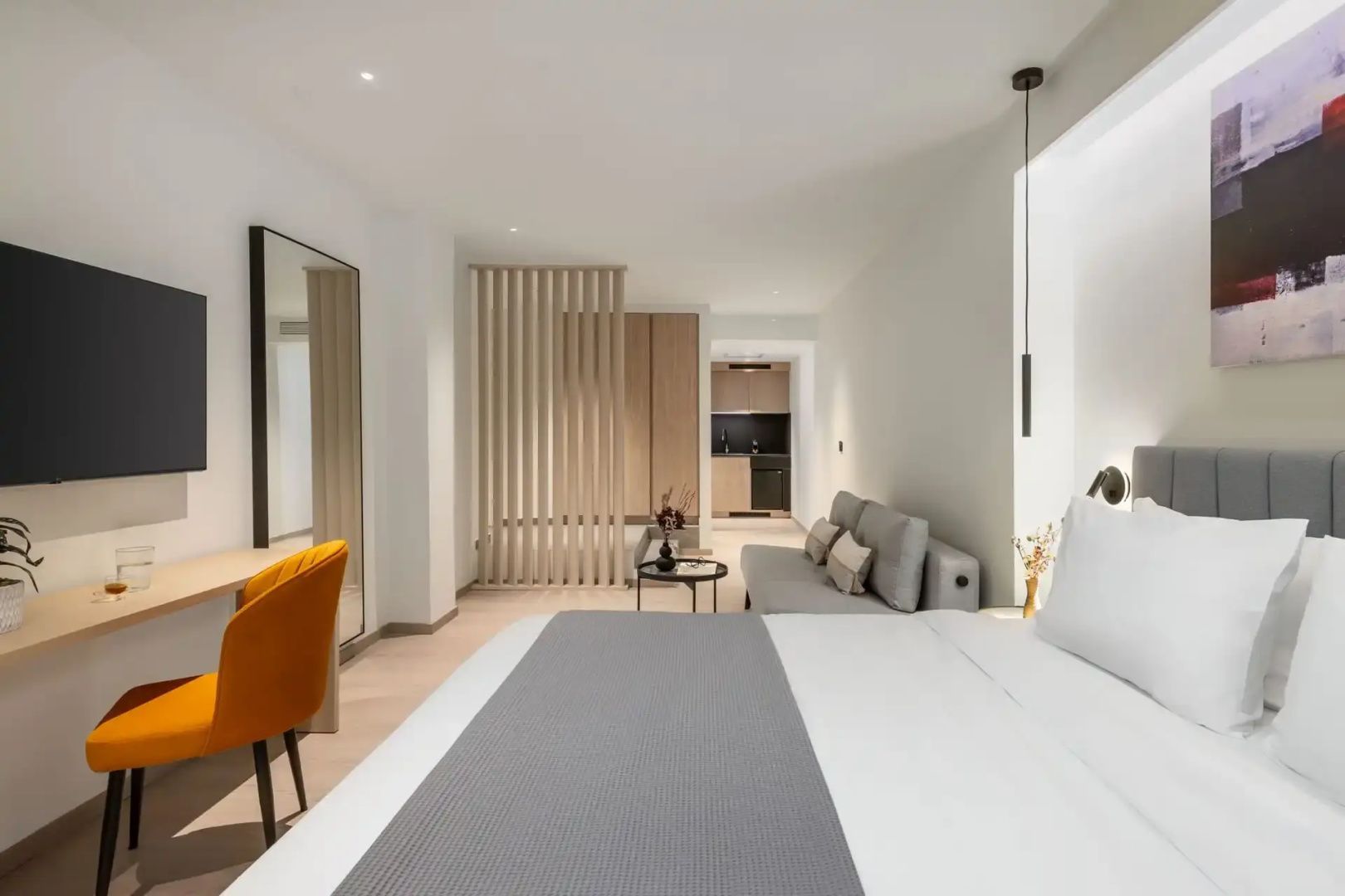 Helles Zimmer mit Doppelbett, grauer Couch, Holzabtrennung und kleiner Küchenzeile.