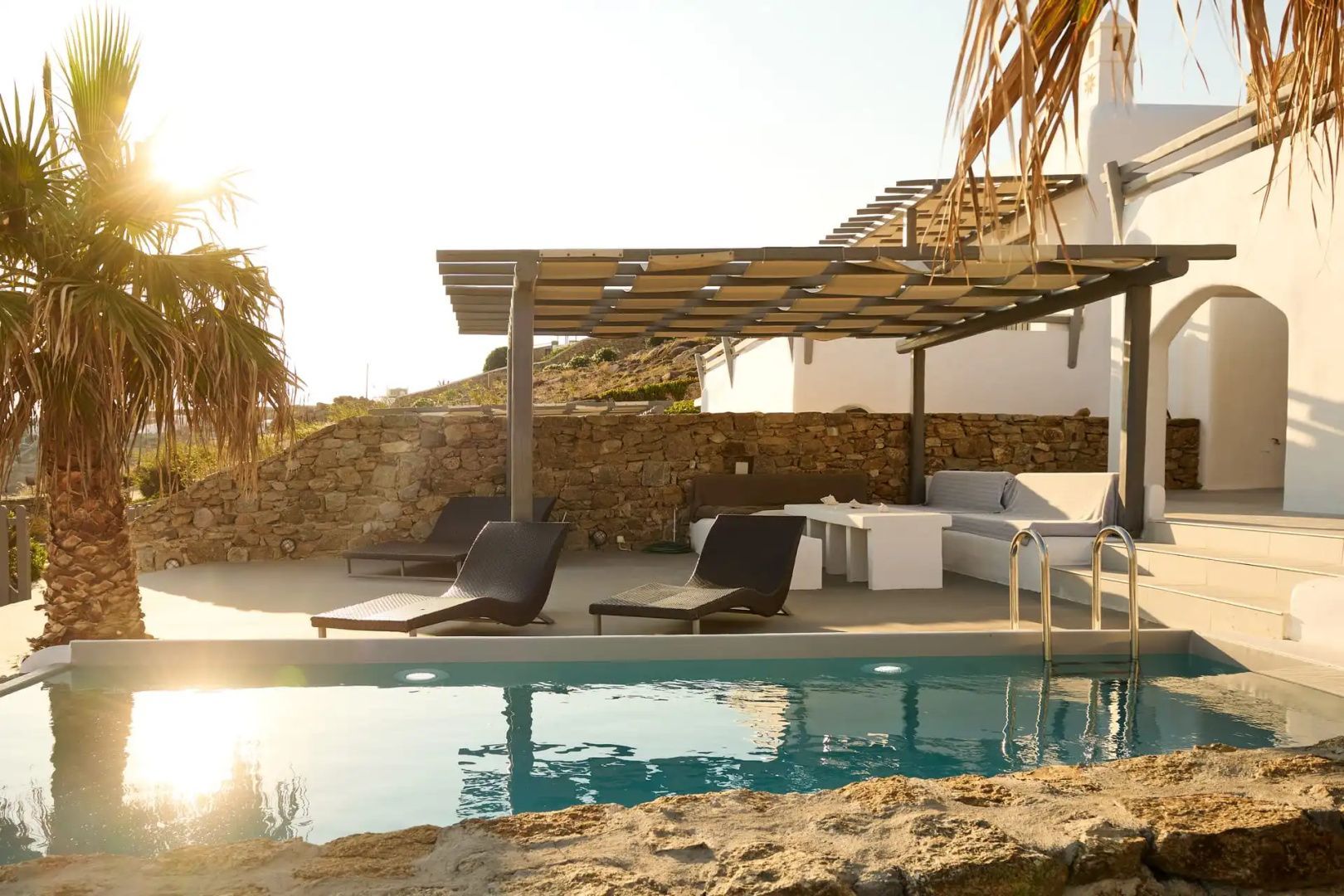Außenpool mit klarem Wasser, Sonnenliegen und überdachter Lounge vor einer Natursteinmauer.