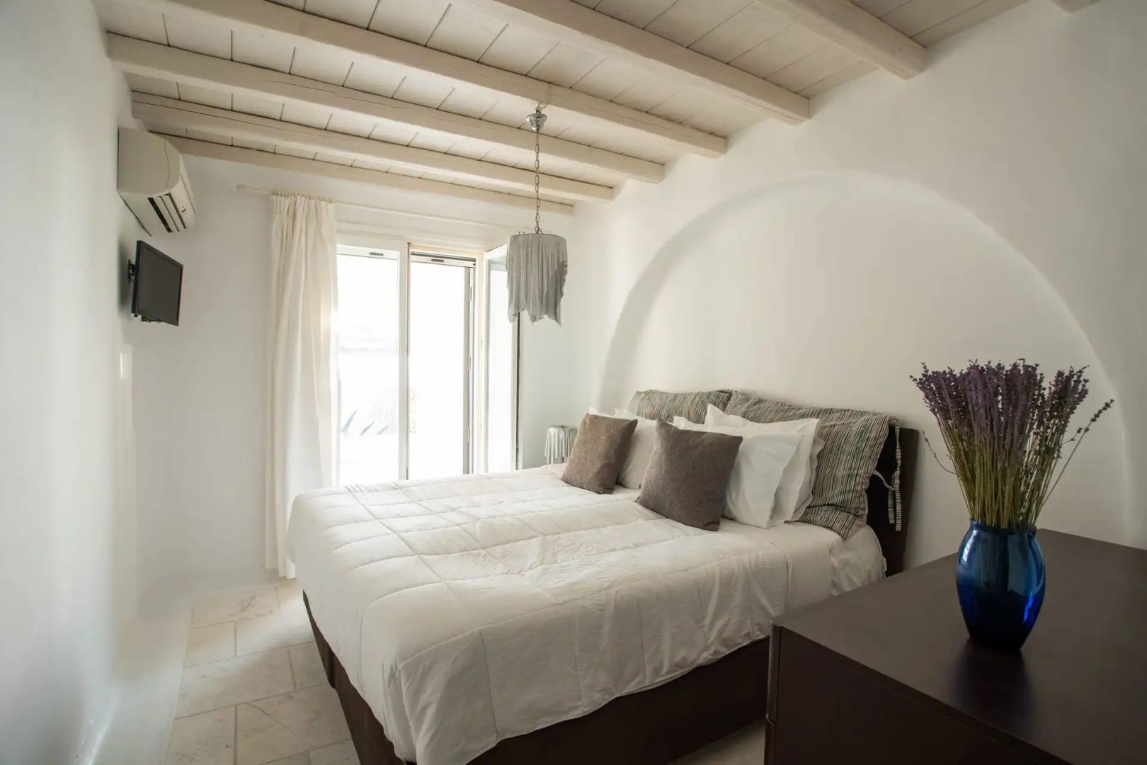 Schlafzimmer mit weißem Doppelbett, Vorhängen am Fenster und einer Decke aus hellen Holzbalken.