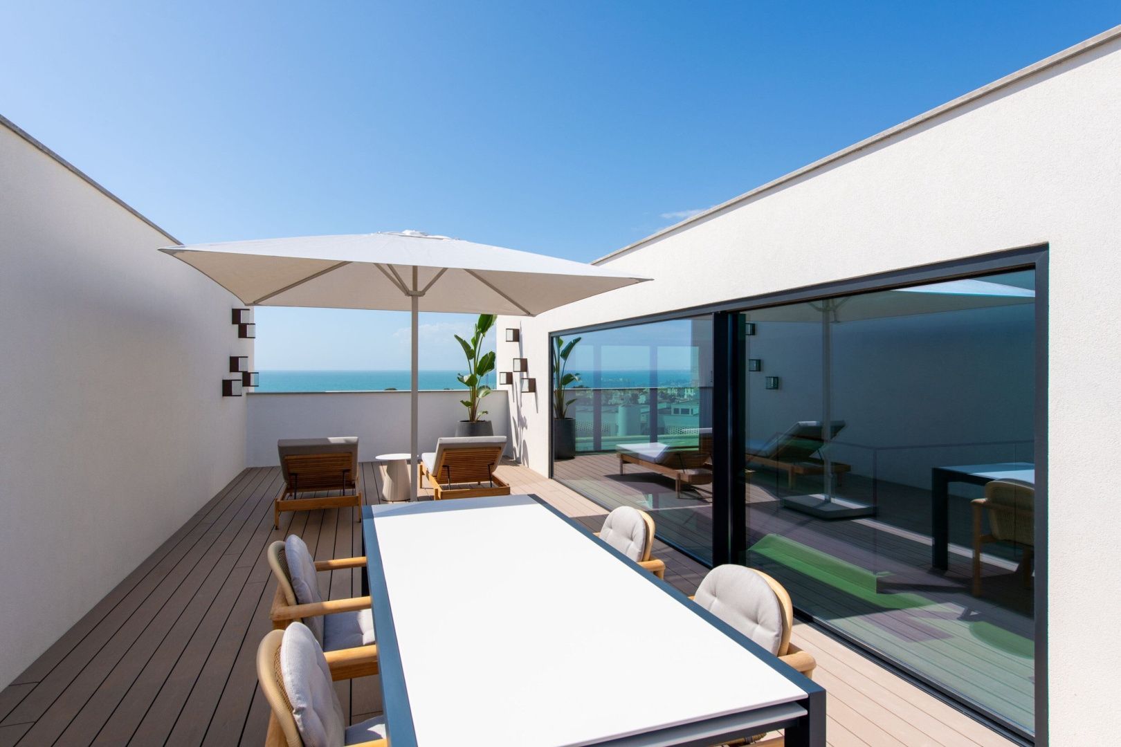 Sonnige Dachterrasse mit Esstisch, Sonnenschirm, Liegestühlen und direktem Blick auf das Meer.