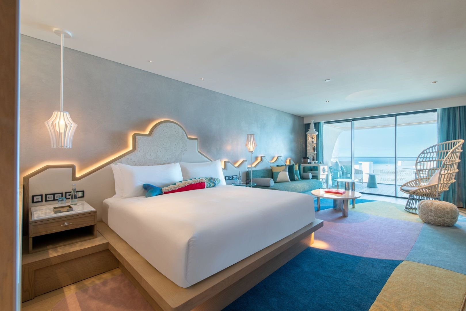 Doppelbett mit beleuchtetem Kopfteil, buntem Teppich und Sofa in einem modern eingerichteten Hotelzimmer.