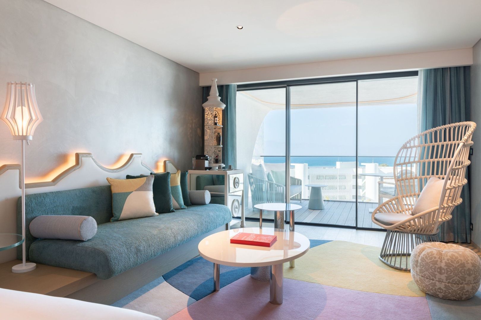 Wohnbereich eines Zimmers mit grünem Sofa, rundem Tisch, buntem Teppich und Blick auf den Balkon.
