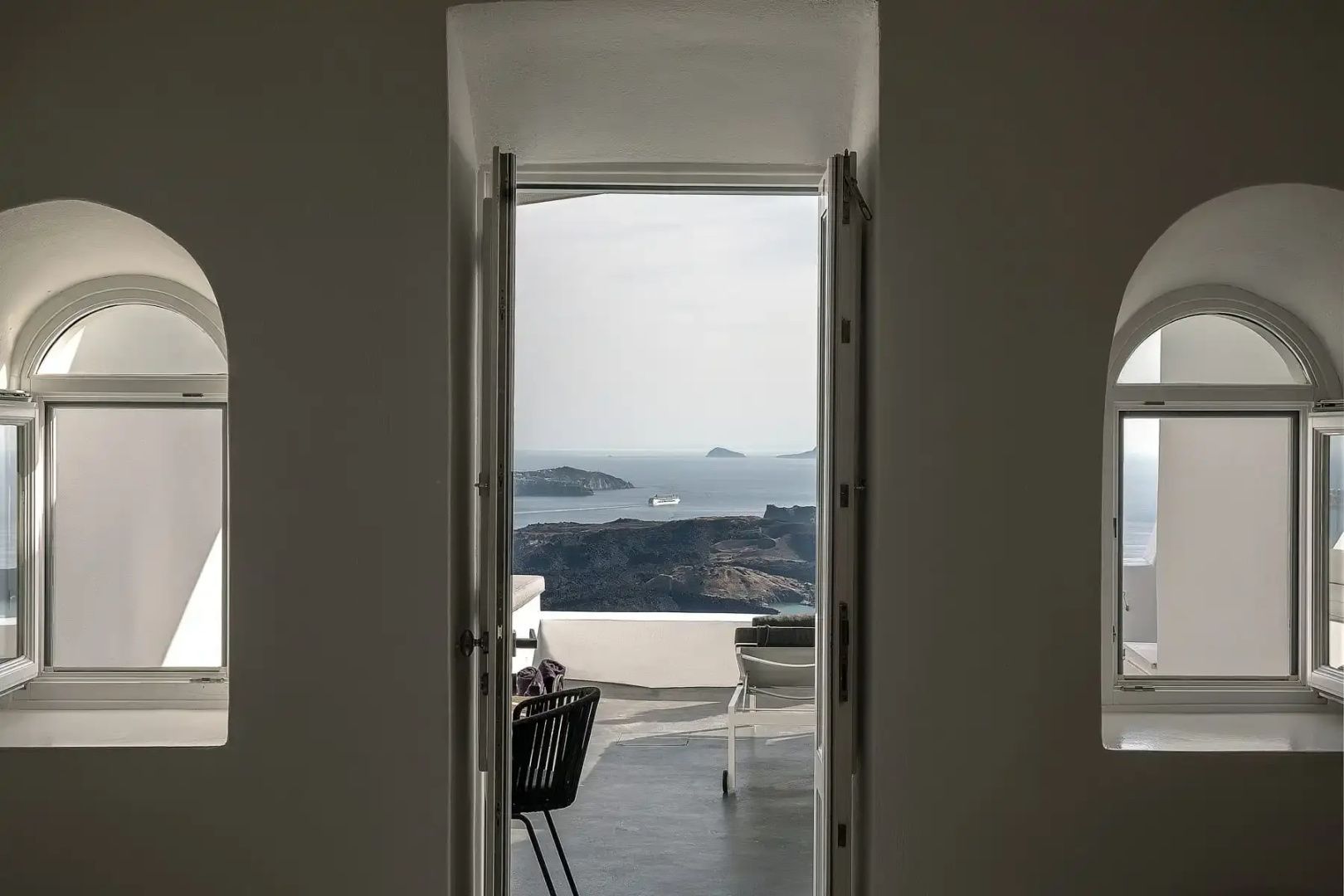 Blick durch ein geöffnetes weißes Fenster auf die Terrasse und das Meer mit Inseln.