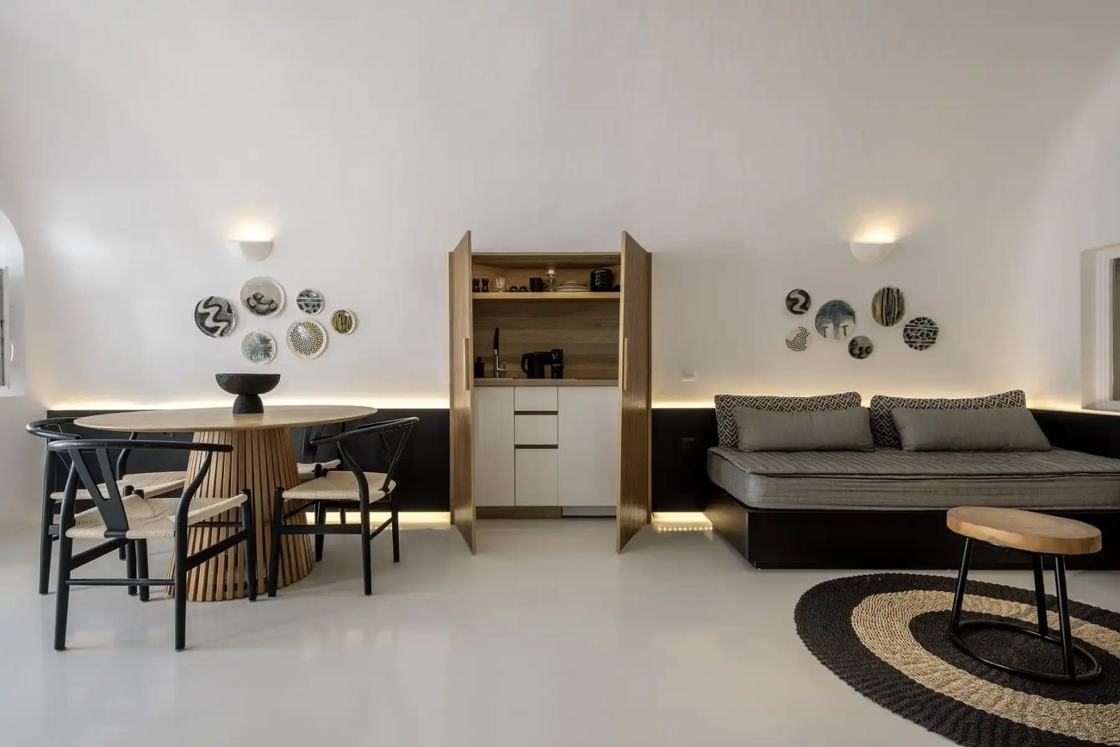 Wohnraum mit rundem Holztisch, grauem Polstersofa und geöffnetem Küchenschrank.