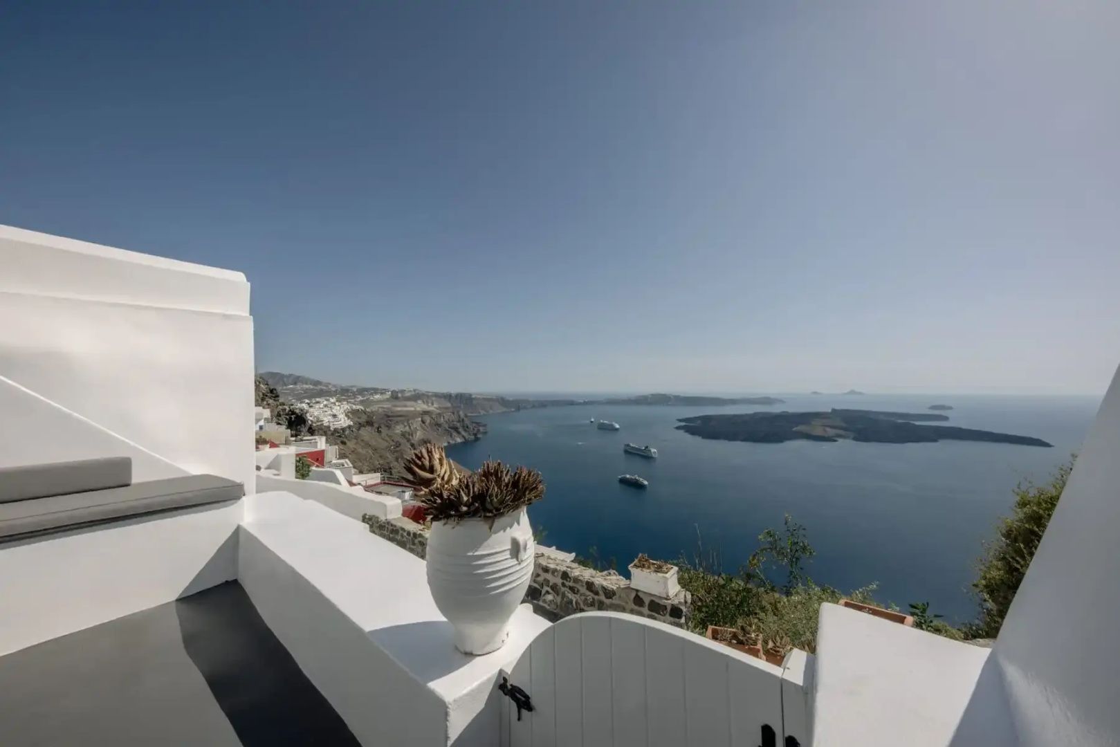 Terrasse mit weißer Tonvase und Blick auf das blaue Meer mit Vulkaninseln und Schiffen.