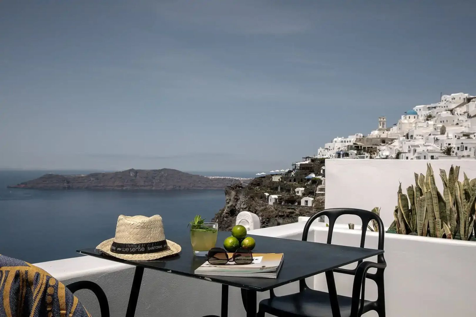 Terrassentisch mit Strohhut und Blick auf weiße Häuser an den Klippen von Santorini.
