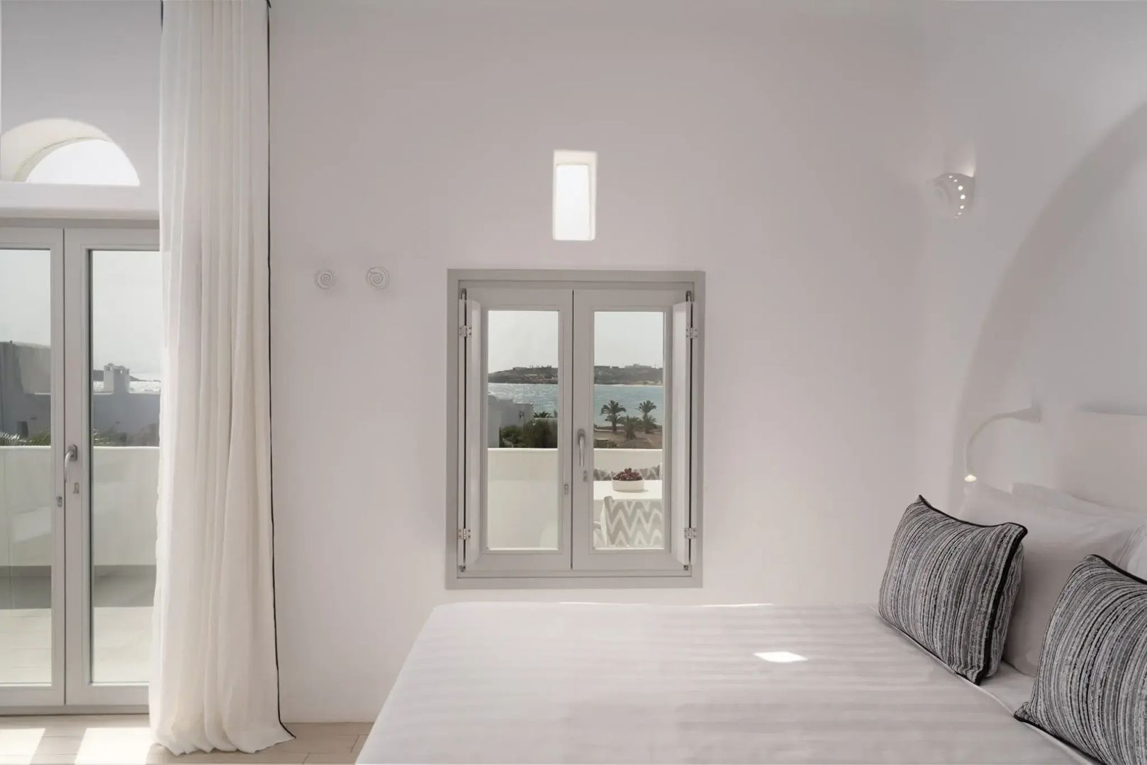 Blick durch ein Fenster im weiß gestalteten Schlafzimmer auf das Meer.