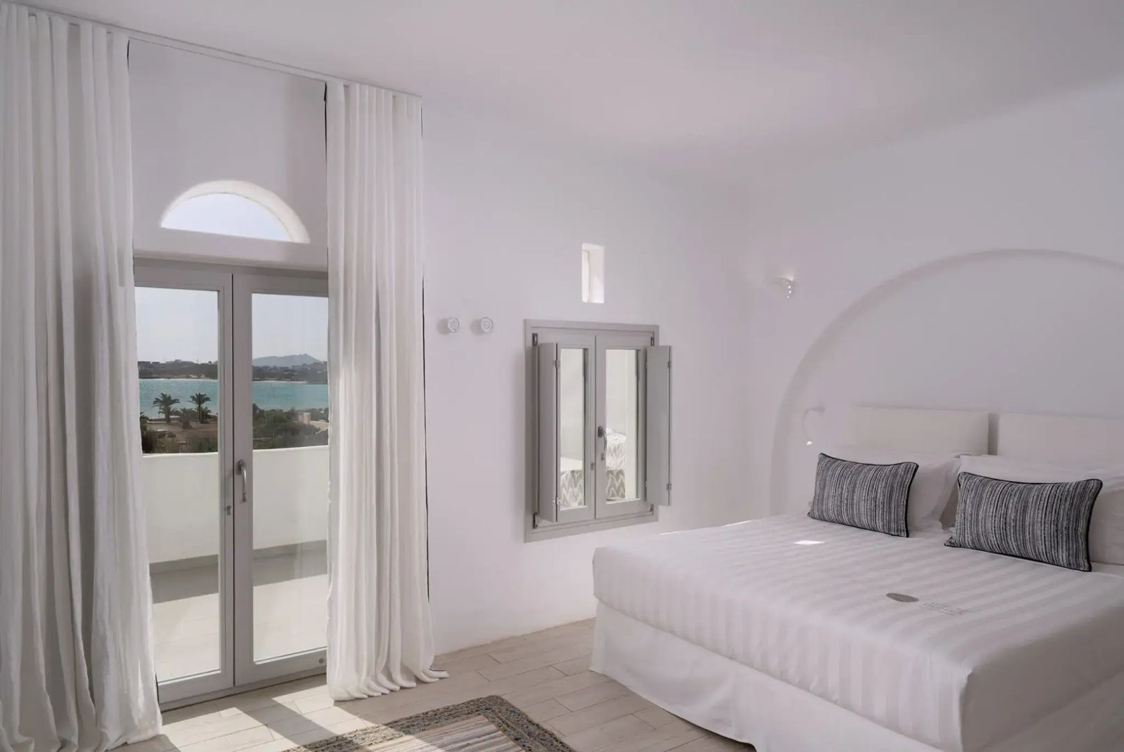 Helles Hotelzimmer in Weiß mit Doppelbett, Vorhängen und einer Balkontür.