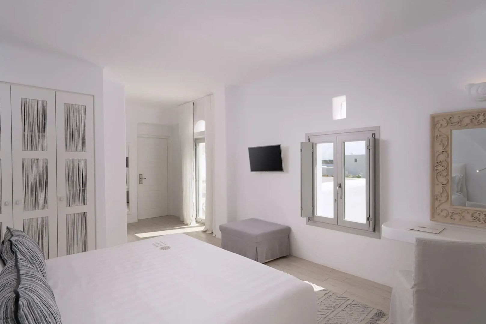Helles Hotelzimmer in Weiß mit Fernseher an der Wand, Fenster und Sitzbank.