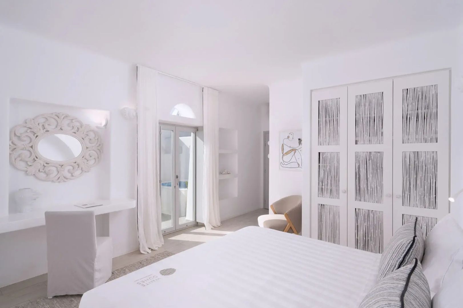 Weißes Hotelzimmer mit Doppelbett, Sessel, weißem Einbauschrank und Balkontür