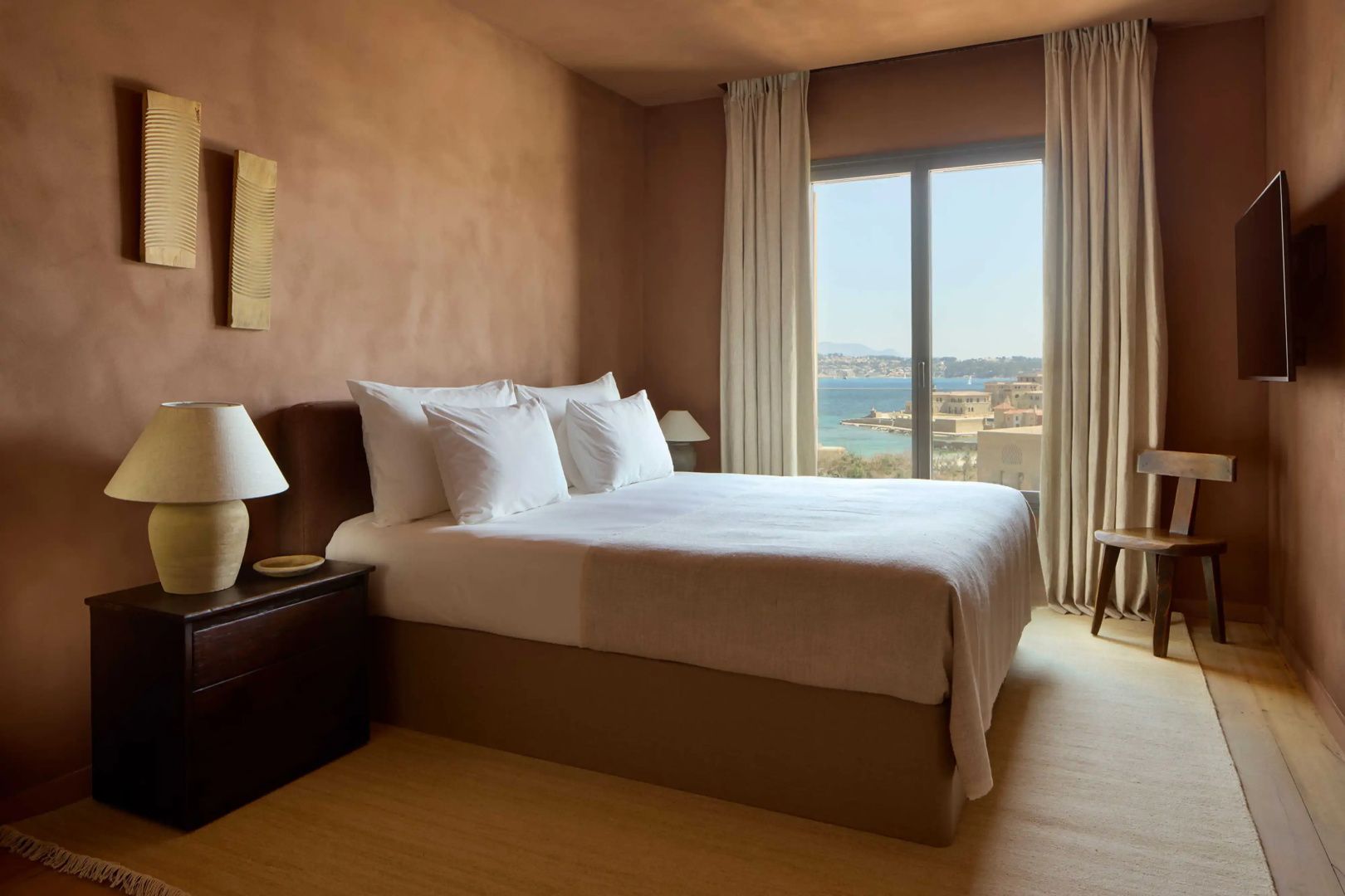 Doppelbett in einem hellen Schlafzimmer mit Blick aus dem Fenster auf das Meer und die Küste.