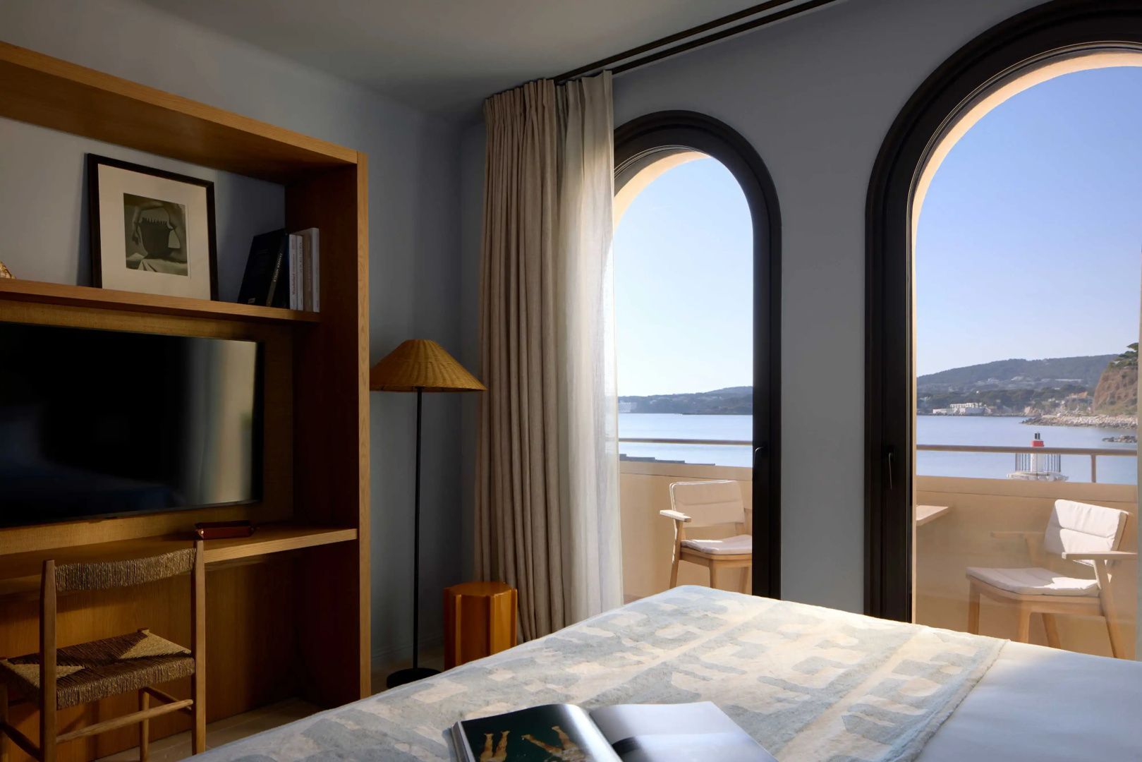 Schlafbereich mit Holzregal, Fernseher und Blick durch Rundbogenfenster auf das Meer.