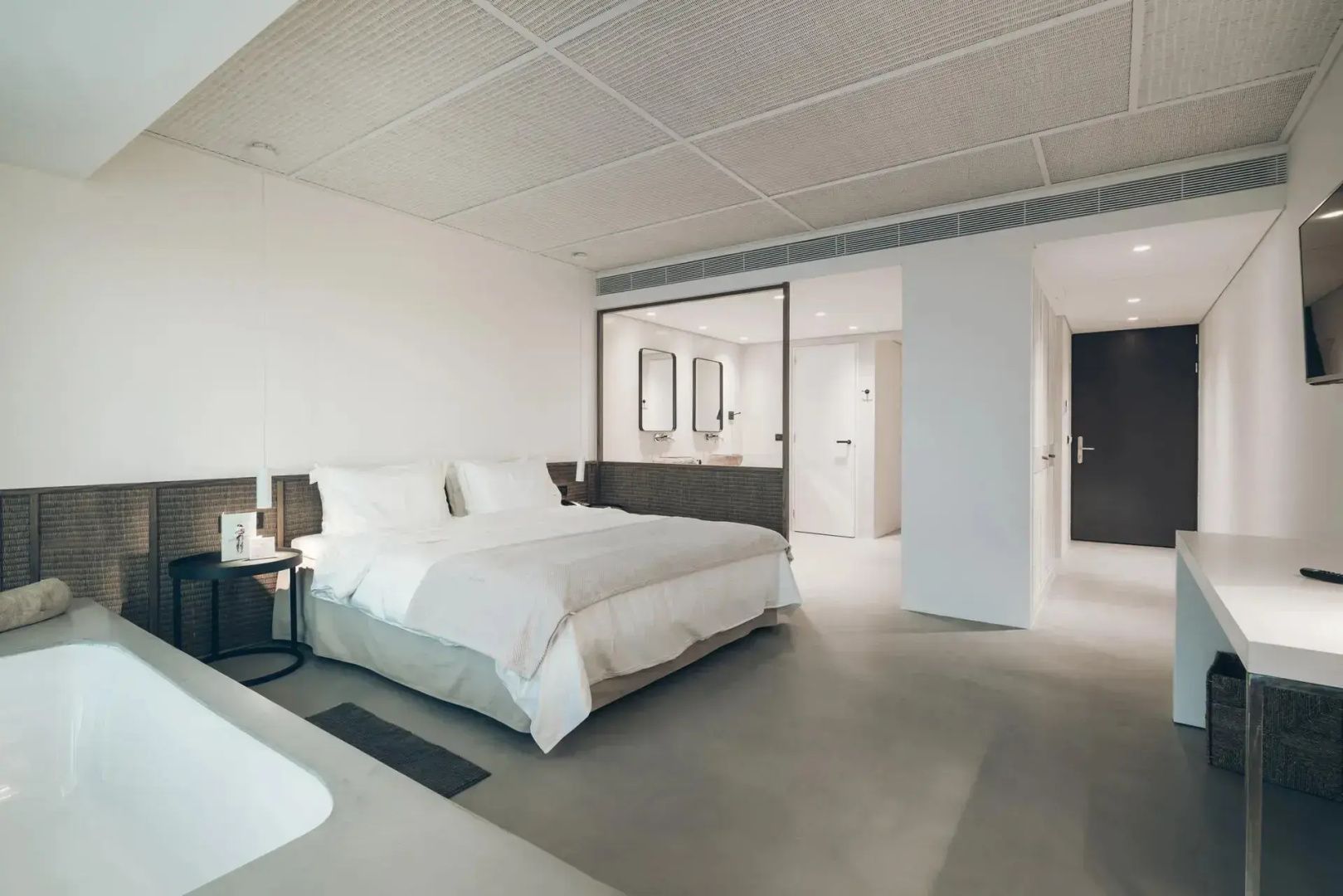 Hell eingerichtete Suite mit weißem Doppelbett, Glaswand zum Badezimmer und hellem Estrichboden.