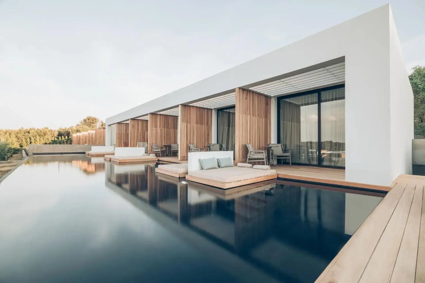Poolbereich mit im Wasser stehenden Liegen und holzverkleideten Terrassen der Suiten.