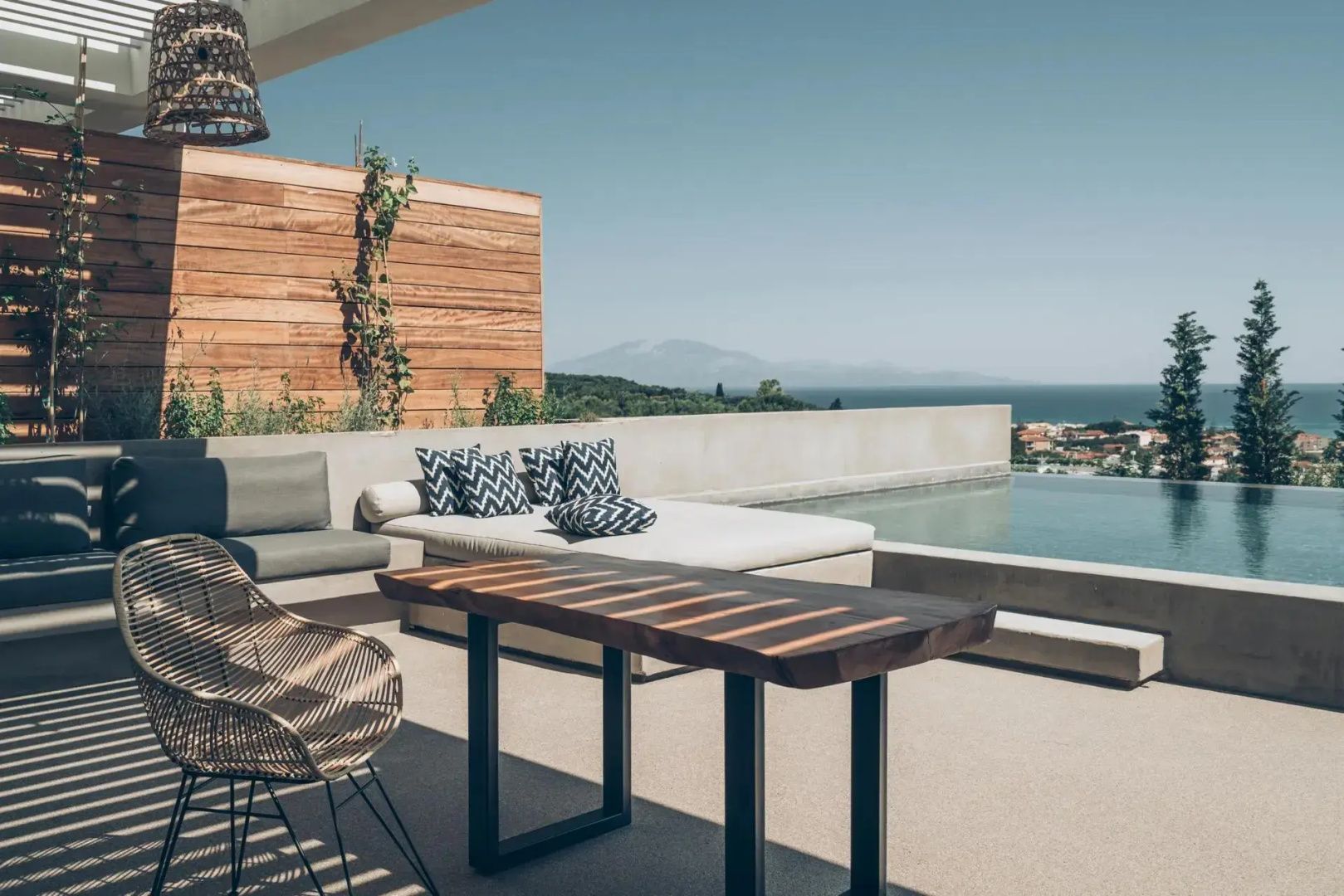 Terrasse mit Holztisch, Liegebett und Blick über einen Pool auf das Meer im Hintergrund.