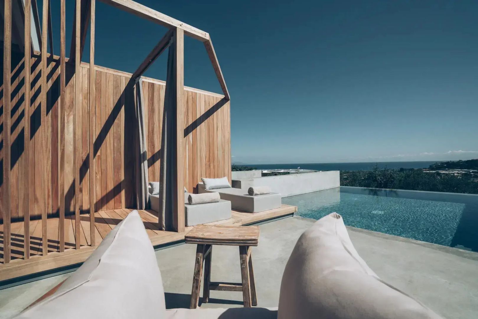 Blick über eine Holzterrasse mit Sonnenliegen und Holzschemel auf einen Infinity-Pool und das Meer.