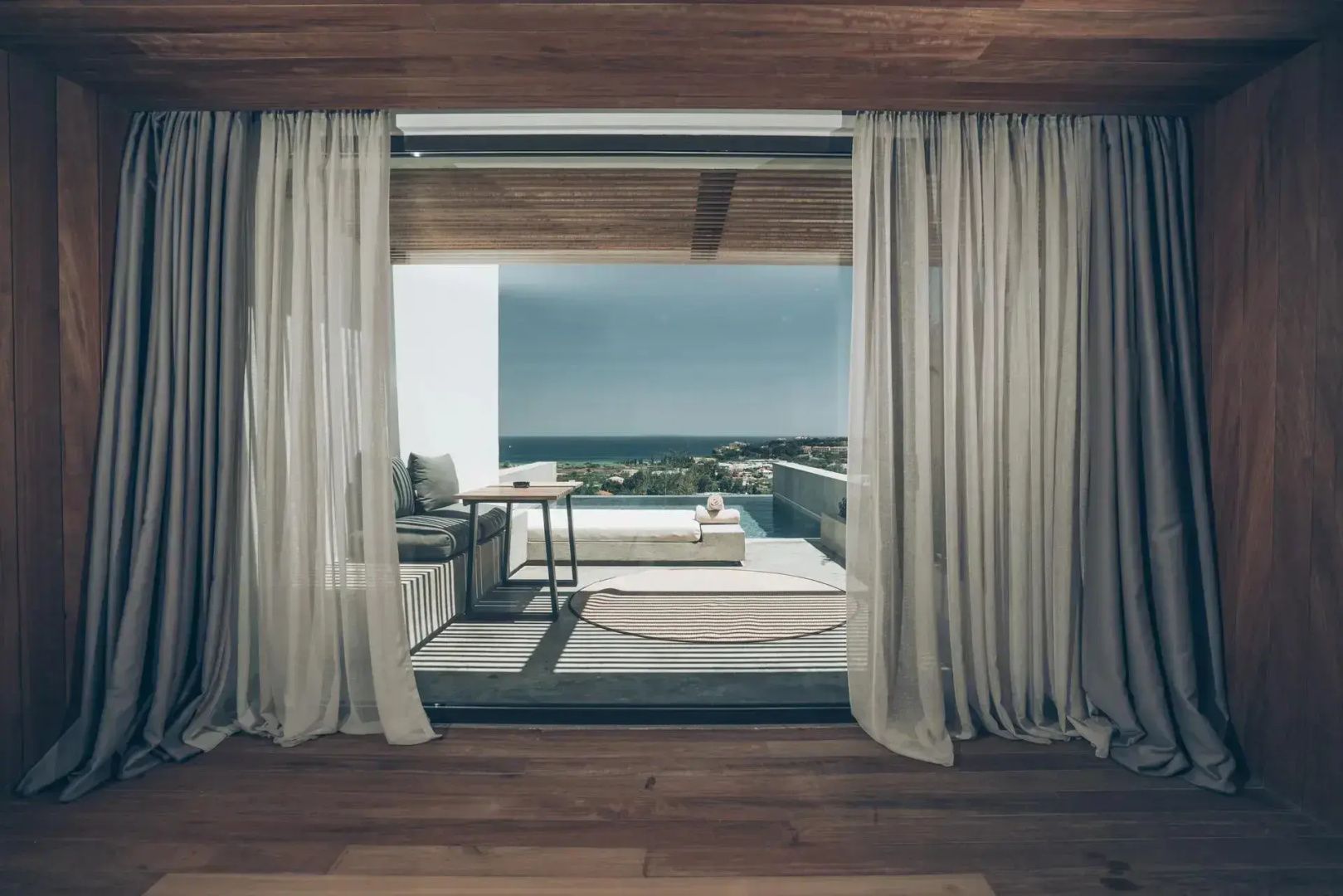 Blick durch geöffnete Balkontüren mit Vorhängen auf eine Terrasse mit Pool und das Meer.