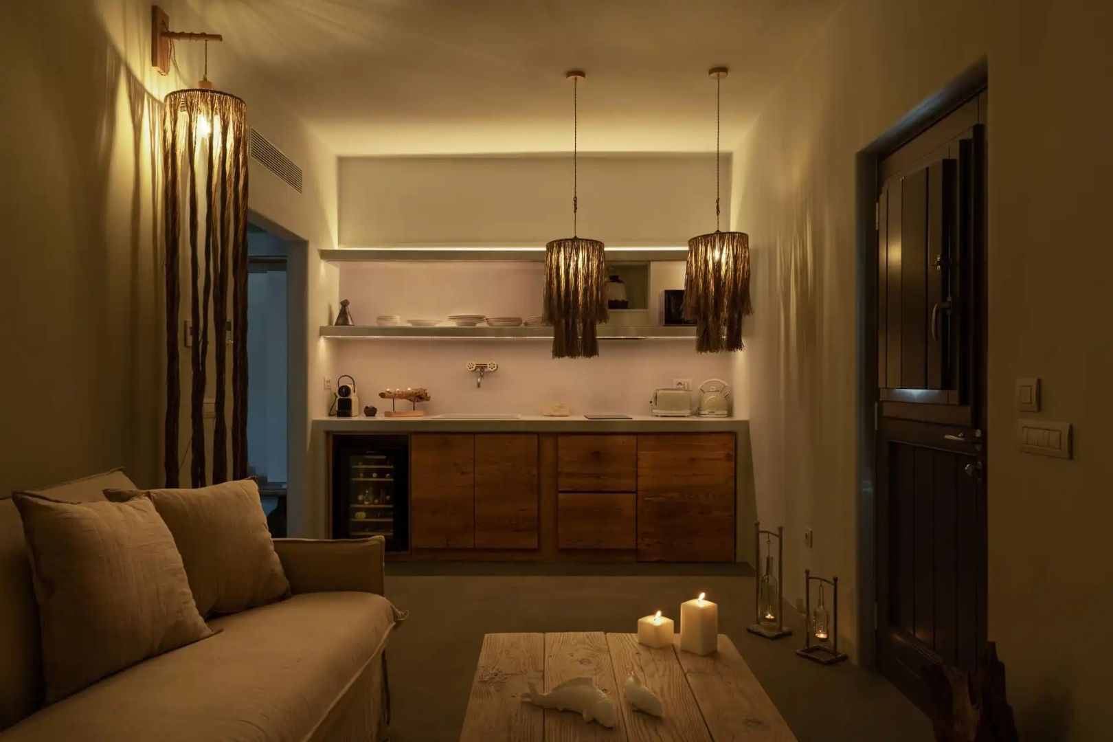 Wohnbereich mit Sofa, Holztisch mit Kerzen, Hängeleuchten und integrierter Küchenzeile im Hintergrund.